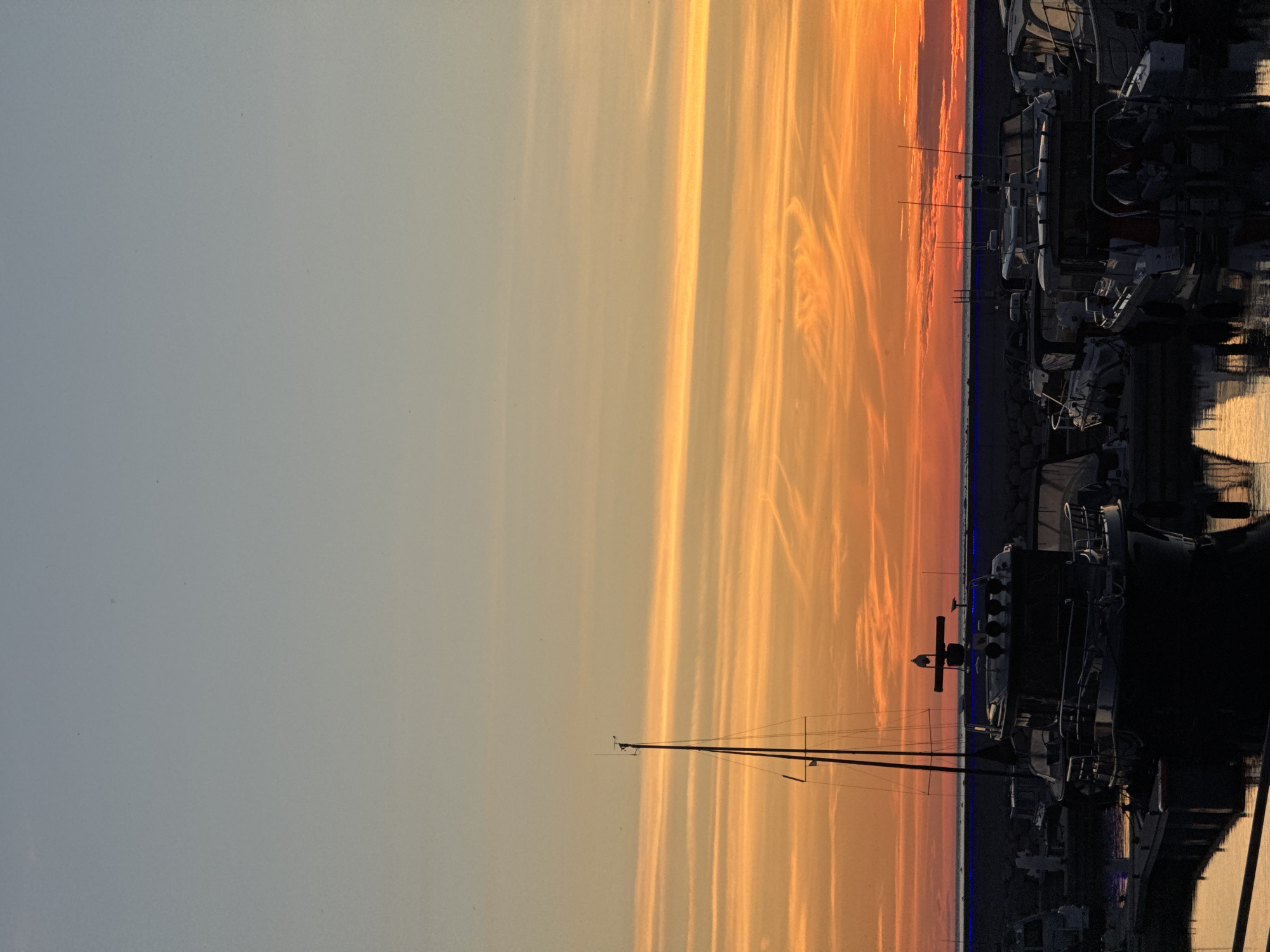 The image depicts a harbor at sunset, with the sky displaying vibrant shades of orange and yellow from the setting sun. Several boats are docked in the foreground, their silhouettes stark against the colorful sky. A sailboat with a tall mast is prominent on the left. In the background, a stone wall separates the harbor from the sea. The water reflects the colors of the sky. The scene appears to be a marina in Tallinn, Estonia, possibly in the Pirita or Old City Harbor area, given the common presence of sailboats and the architectural style of some of the boats which could suggest a Northern European marina. There are no people visible in the image.