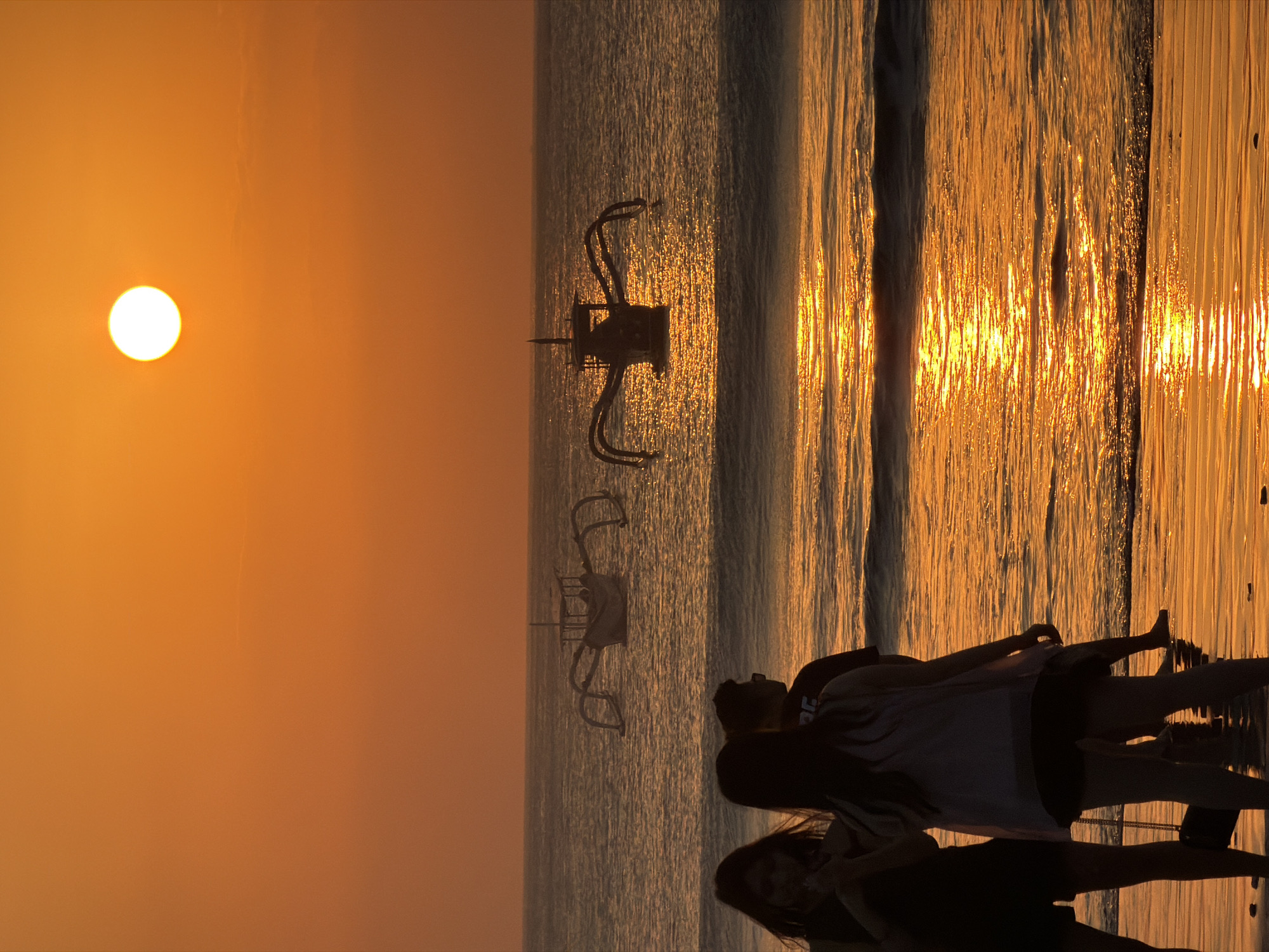 The image captures a breathtaking sunset over the ocean at Kuta Selatan, Indonesia. The sky is a vibrant gradient of orange and yellow, with the sun a bright white orb positioned in the upper right quadrant. Two traditional Balinese fishing boats, known as "jukung," are silhouetted against the shimmering water. These boats are characterized by their outriggers, which extend outwards from the main hull, giving them a distinctive appearance.
In the foreground, three individuals stand on the wet sand of the beach, their figures silhouetted against the sunset. One person, appearing to be a woman, is wearing dark clothing and shorts. Next to her, another person, also appearing to be a woman, is wearing a light-colored top over dark shorts. The third person, in the background, is a man wearing a dark t-shirt. Their backs are to the camera, and they appear to be observing the sunset.
The ocean is calm, with gentle waves lapping at the shore. The setting sun casts a golden reflection on the water's surface, creating a beautiful shimmering effect. The wet sand in the foreground also reflects the warm hues of the sunset, adding to the overall serene and picturesque atmosphere of the scene. The image evokes a sense of tranquility and natural beauty, typical of sunsets in this popular Indonesian tourist destination.