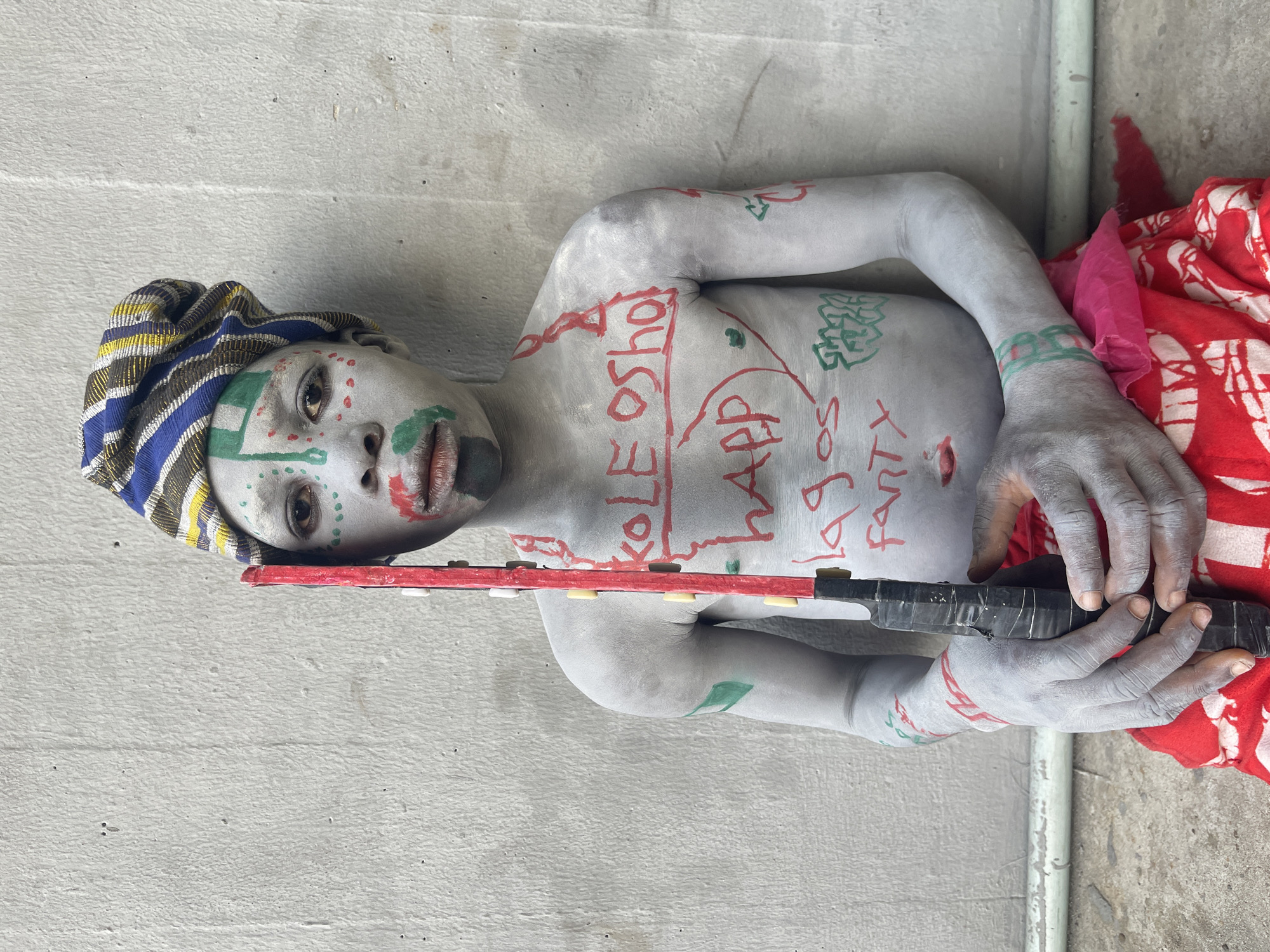 A person lies on their side against a textured gray wall, possibly concrete. The person’s skin is painted gray and adorned with red and green markings. Red writing on the torso reads "KOLE OSHO HAPPY LAGOS PARTY". The person wears a striped head covering and holds a black object with a red extension across their body. A red and white patterned fabric is draped behind the person. The location is the city of Lagos, Nigeria.