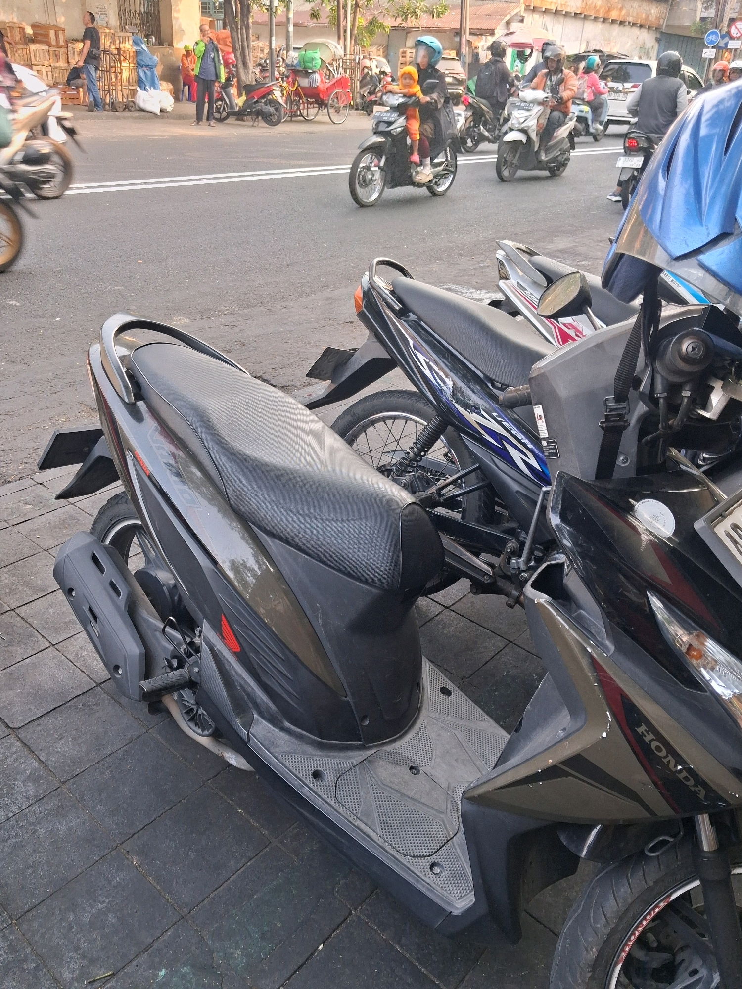 The image depicts a street scene in Surakarta, Indonesia, with a focus on motorcycles and pedestrian activity. In the foreground, a dark gray Honda scooter is parked on a tiled sidewalk. Its seat and exhaust pipe are clearly visible. Behind it, another motorcycle, primarily black with blue accents and the word "YAMAHA" visible, is also parked. A bright blue motorcycle helmet is positioned in the upper right of the frame, partially obscuring a portion of the blue and black Yamaha.
In the background, the street is bustling with activity. Several motorcycles are on the road, some with riders. One scooter in the middle ground, ridden by a person wearing a helmet, appears to be in motion. Further in the background, a red rickshaw is visible, along with other parked motorcycles and some people. Buildings line the street, and a stack of wooden crates is present on the left side. The environment suggests a typical urban setting in Surakarta, likely a commercial or high-traffic area. The lighting indicates it is daytime, possibly late afternoon or early morning, given the quality of the light.