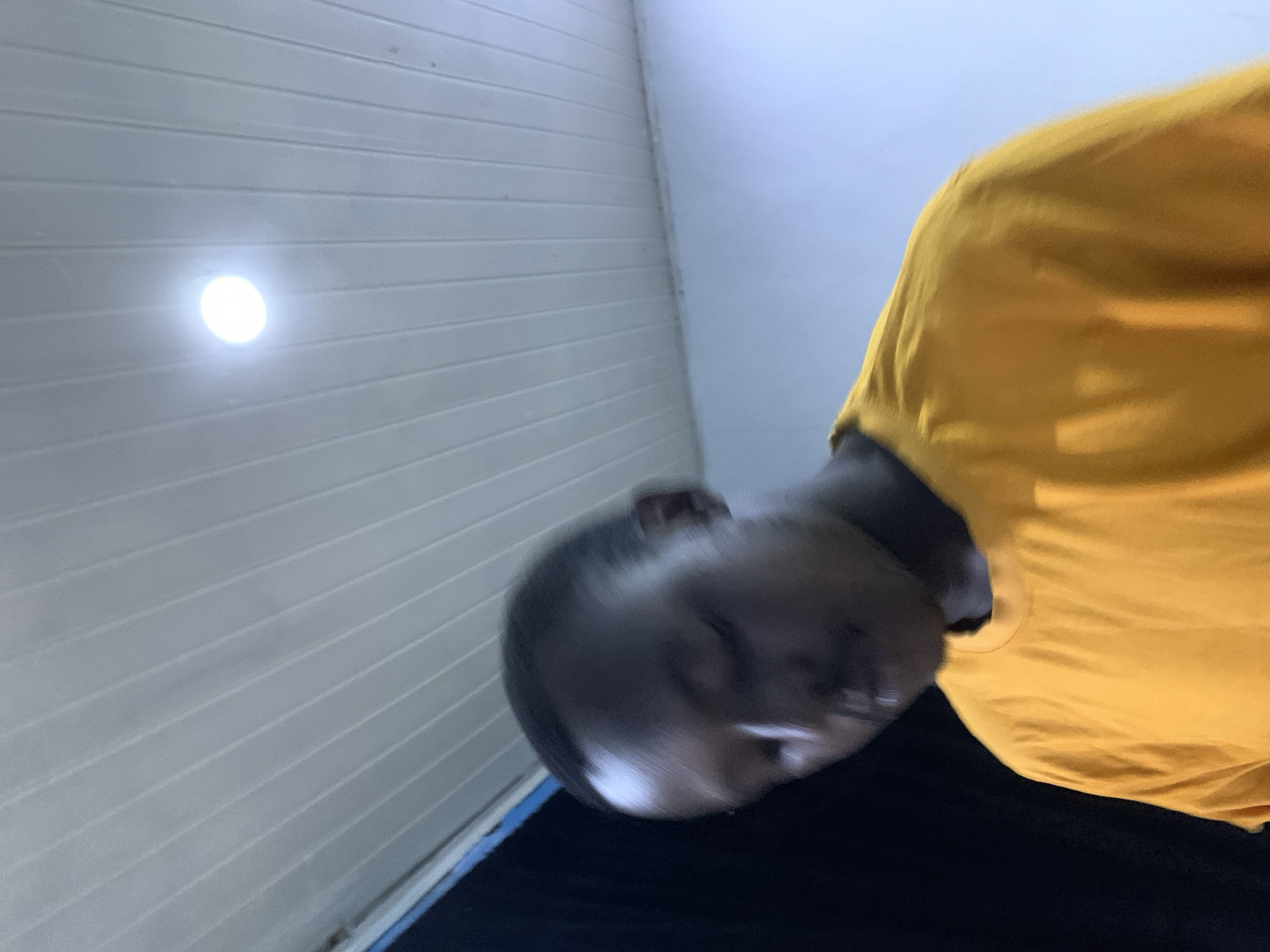 The image presents a low-angle perspective of a person, likely taken indoors or under a covered structure in Jos, Nigeria. The main subject is a person, whose head and upper torso are visible, positioned in the lower-left and central parts of the frame. The person's head is tilted and appears motion-blurred, making their facial features and expressions indistinct. They are wearing a bright yellow short-sleeved t-shirt and dark trousers, partially visible at the bottom of the frame.
The background consists primarily of a white or light-gray paneled ceiling, characterized by numerous horizontal grooves or lines. A distinct, bright, circular artificial light source is embedded in this ceiling, located in the upper-left area of the image. Towards the upper-right, a pale blue or light-gray wall or surface is visible, meeting the paneled ceiling. The unusual upward-looking angle suggests the photograph was taken from a very low vantage point. There is no discernible activity beyond the person's presence and posture. No visible text is present in the image.