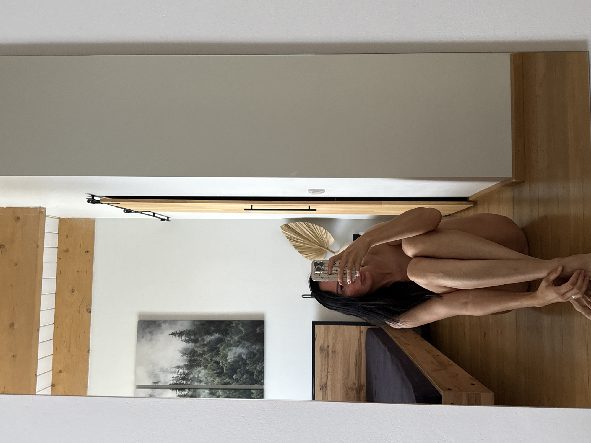 A woman is seated on the floor in front of a mirror, taking a selfie with her phone. She is wearing no clothes and her legs are tucked underneath her. A bed frame made of light-colored wood is visible behind her to the left, with a dark mattress. A large, framed artwork of a forest hangs on the wall above the bed. A wooden sliding door is visible to the right of the frame. The floor is made of polished wood. The room appears to be a bedroom. The location is not specified in the image or the prompt.