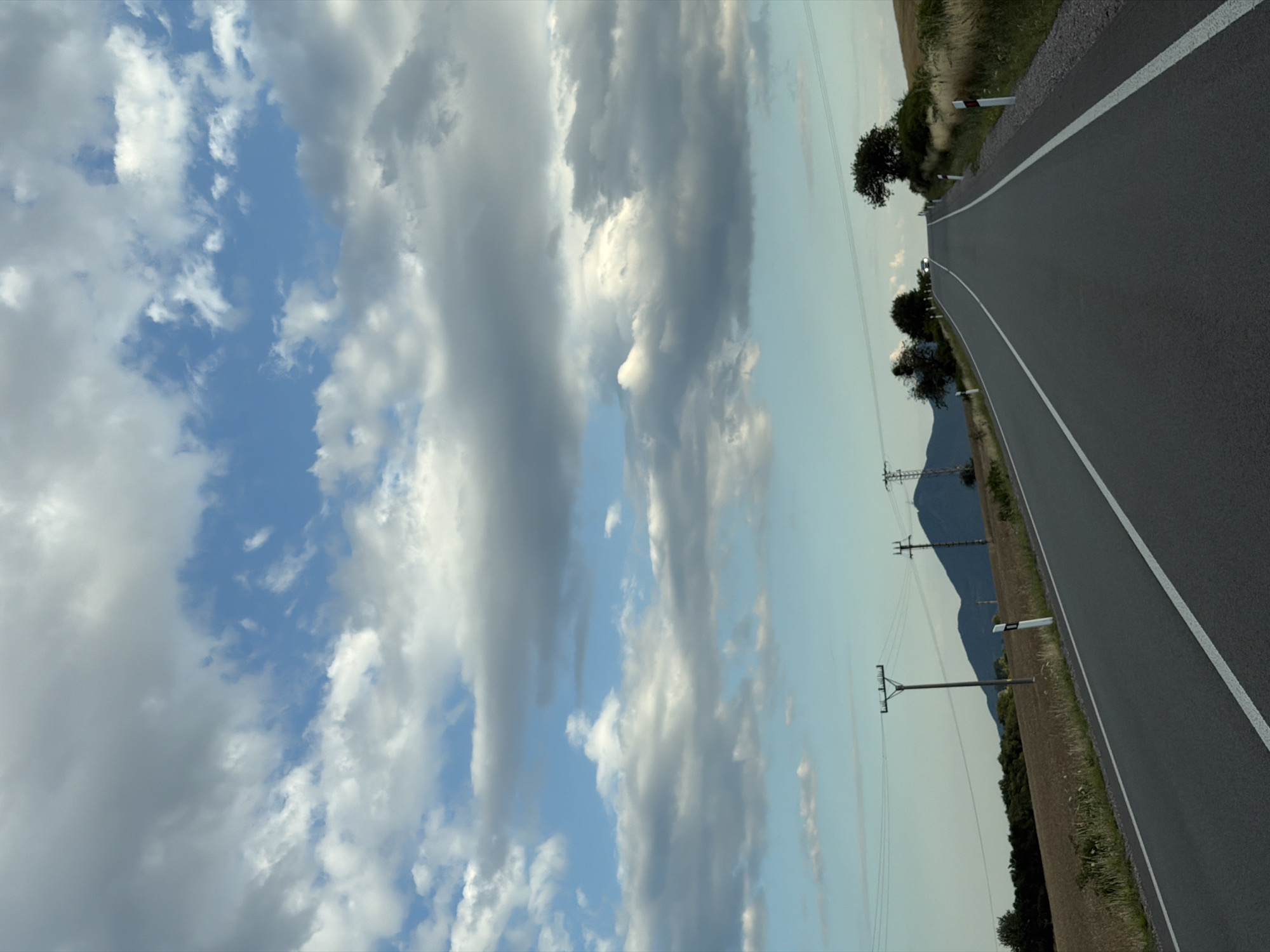 The image shows a view of a road in Luhyňa, Slovakia, leading into a gently sloping hill. The road is paved with asphalt and has white lines marking the lanes. The sky is partly cloudy with a mix of blue sky and grey and white clouds. On either side of the road, there are fields of dry grass and some scattered trees. In the distance, there are mountains and power lines. There is a single car visible on the road in the far distance, its headlights on. There are no people visible in the image. The scene appears to be a rural landscape.