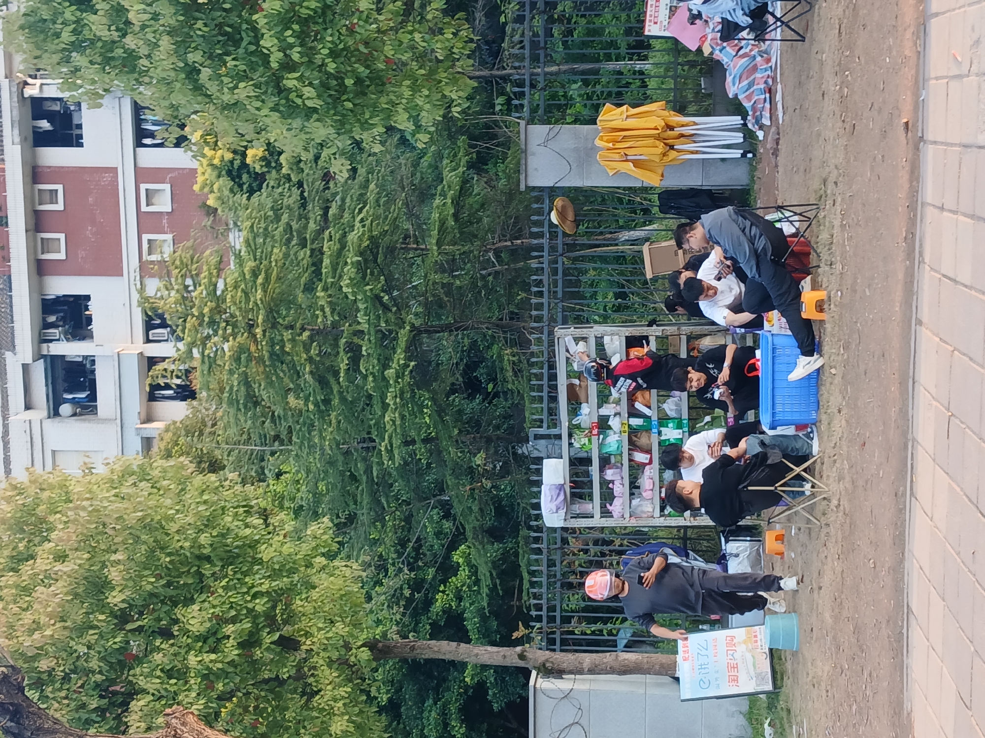 The image depicts a street scene in Shapingba District, China. Several people are seated in a row on chairs, apparently receiving pedicures from individuals kneeling before them. A metal shelving unit is positioned behind the seated people, holding various items, including shoes, bags, and packaged goods. Behind the group is a metal fence with trees and foliage behind it. To the left of the group is a building. A sign on a blue container is visible in the foreground. An individual in a gray outfit and orange helmet is walking in the foreground. Other items visible include folded yellow tents and assorted objects. The setting appears to be outdoors, on a paved walkway or street.