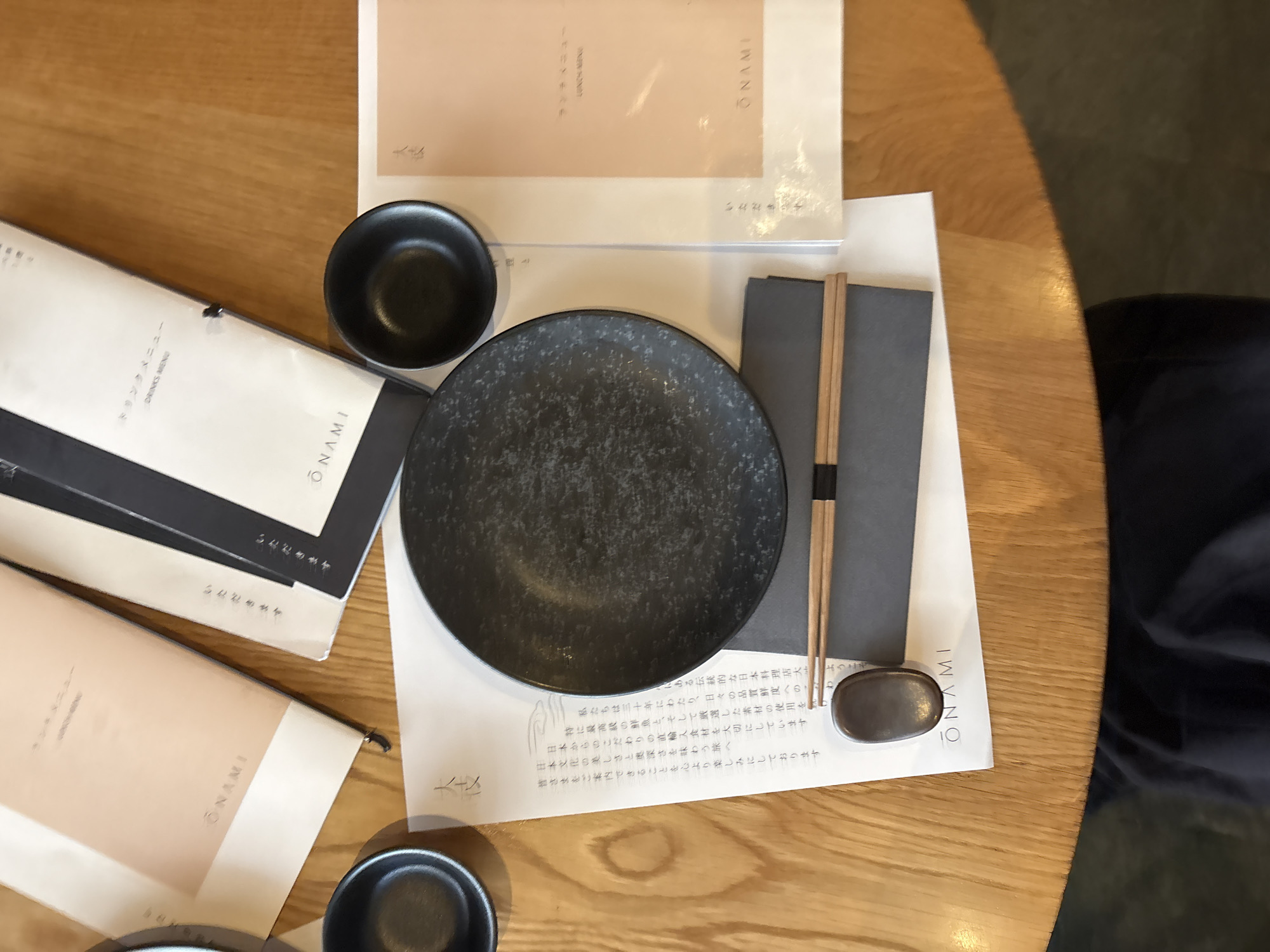 The image shows an overhead view of a table setting at a restaurant. The table appears to be made of light-colored wood. Several items are arranged on the table, including a black speckled plate, two small black bowls, a pair of chopsticks resting on a gray chopstick rest on top of a gray placemat, and a menu. The menu has Japanese text, and the name "ONAMI" is printed on it. There's another menu item and a placemat with the same name on it. The setting suggests a Japanese restaurant. A chair and floor are visible in the background. The scene lacks information to identify its location in Tel-Aviv, Israel.