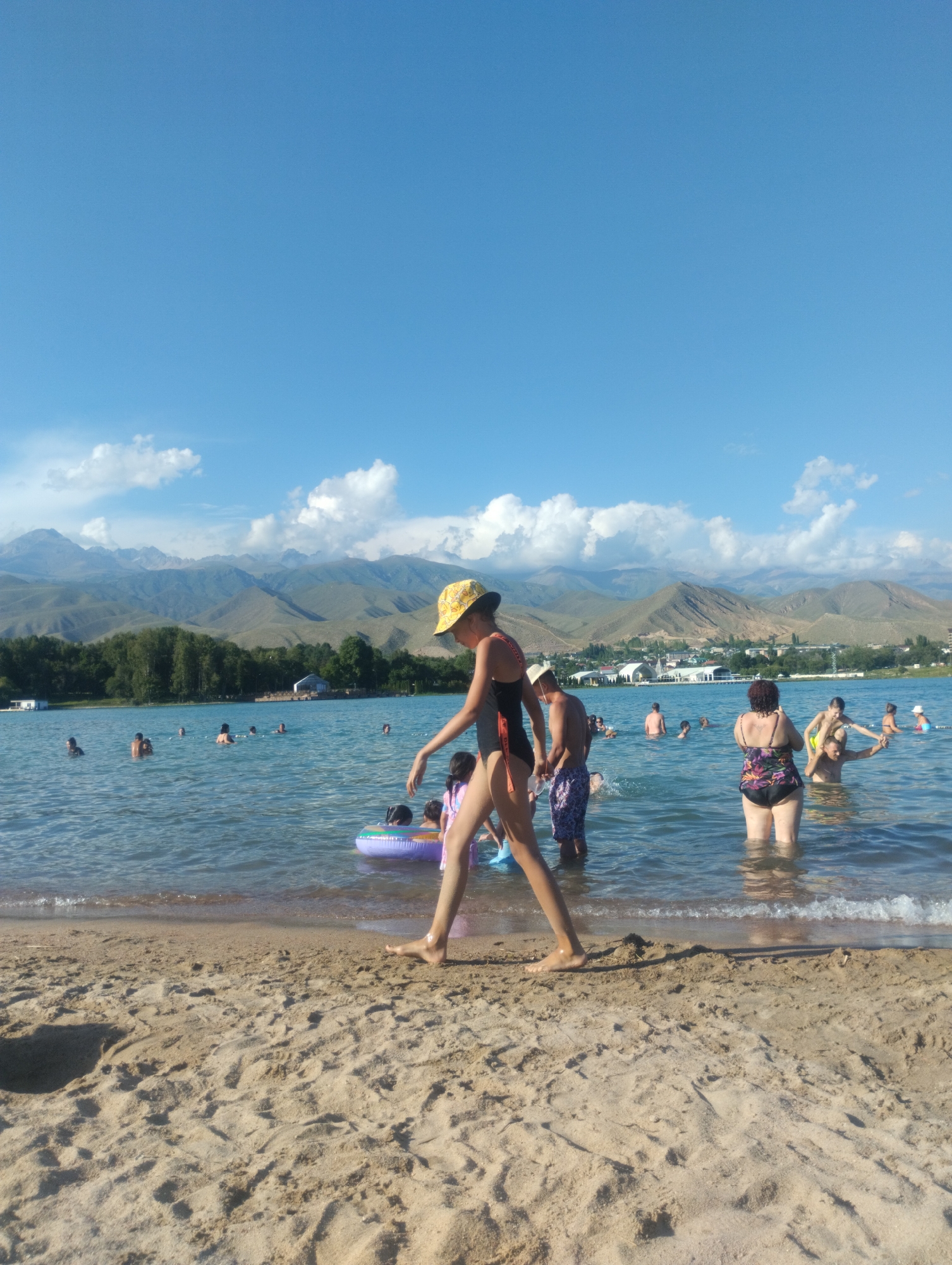 The image depicts a crowded beach scene on a sunny day at Lake Issyk-Kul, in Cholpon-Ata, Kyrgyzstan. People of various ages are enjoying the water, with some swimming and others wading near the shore. In the foreground, a young girl in a yellow hat and black swimsuit walks along the sandy beach. Several other individuals are visible in the water, some with inflatable toys. The background reveals a picturesque landscape with lush green trees, buildings along the shoreline, and distant mountains under a clear blue sky with scattered white clouds.