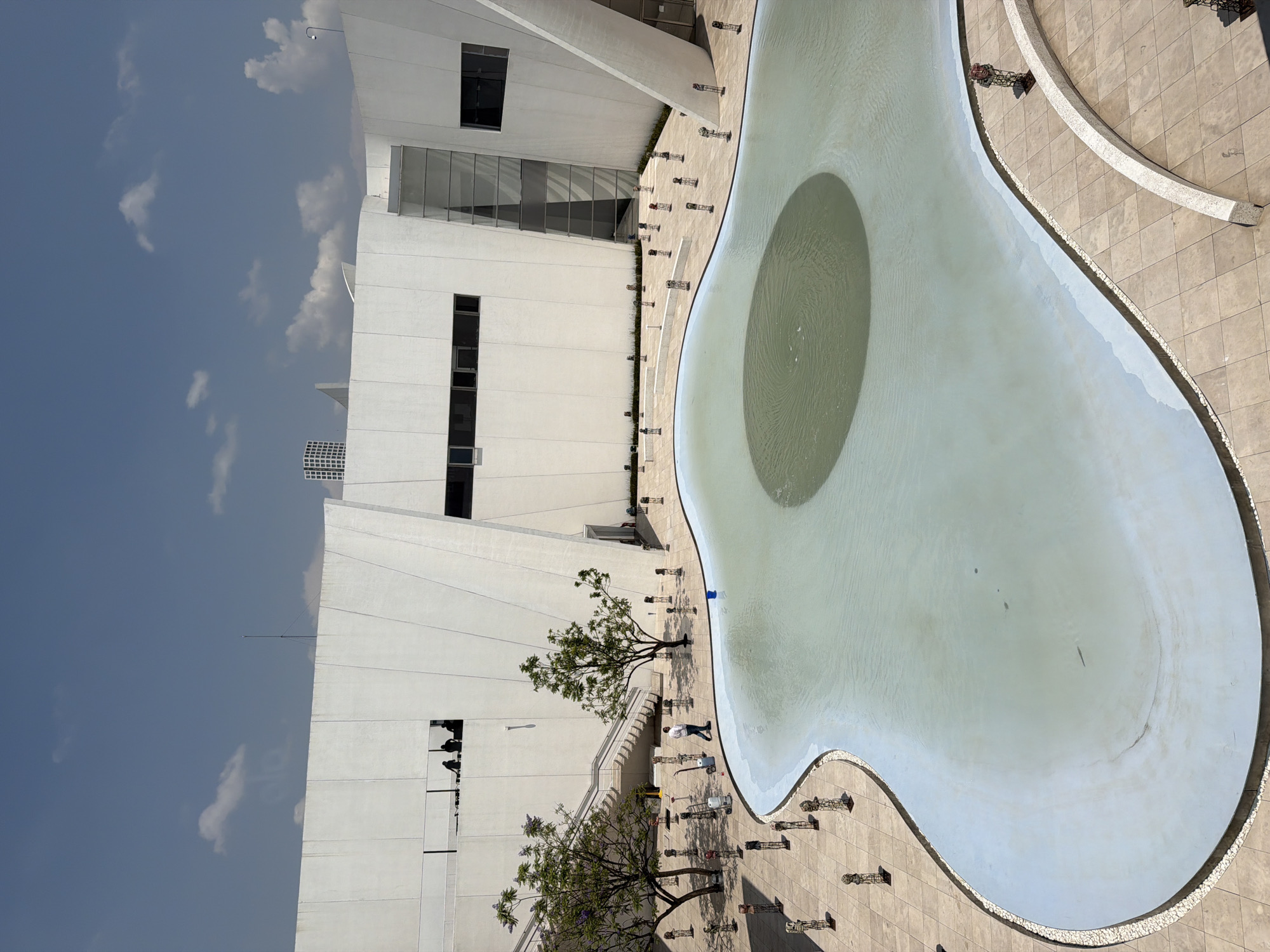 The image depicts a building complex with a large, irregularly shaped pool in San Bernardino Tlaxcalancingo, Mexico. The building is a modern, multi-story white structure with windows and geometric architectural elements. Adjacent to the building, there is a pool with a round darker spot in the middle. There are various decorative structures placed along the pool's edge, resembling ornate lamps or stands. Two trees are situated near the building. People are visible around the pool and the area where the trees are planted. The sky is blue with a few clouds. The ground around the pool and the building is tiled with a light-colored stone.