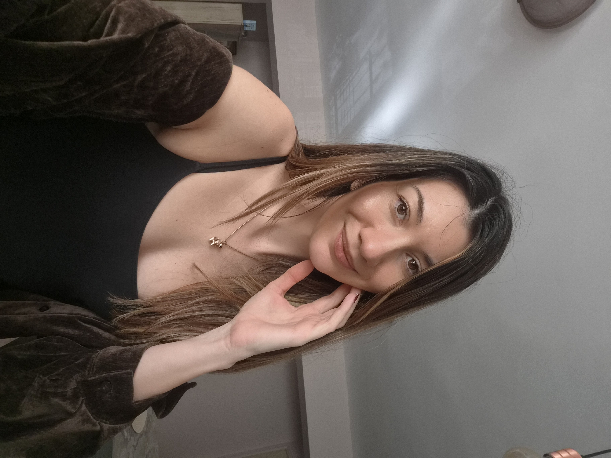 A woman is depicted in a medium close-up shot, looking directly at the camera with a gentle smile. She has long, straight to slightly wavy brown hair with lighter highlights, which cascades around her face and over her shoulders. She is wearing a black top or camisole and a dark brown, possibly velvet or corduroy, jacket or shirt draped over her. A delicate gold-colored necklace with a small animal-shaped pendant (resembling a bear) hangs around her neck. Her left hand, with nails painted a dark shade, is softly placed against her chin.
The background consists of plain, light-colored walls, indicating an indoor setting, likely a modern room or home. Subtle light variations or reflections are visible on the wall surfaces. The lighting is soft and even, typical of indoor illumination. The woman's expression is relaxed and pleasant. The original image appears to be rotated clockwise. No visible text is present. The location context is Plovdiv, Bulgaria.