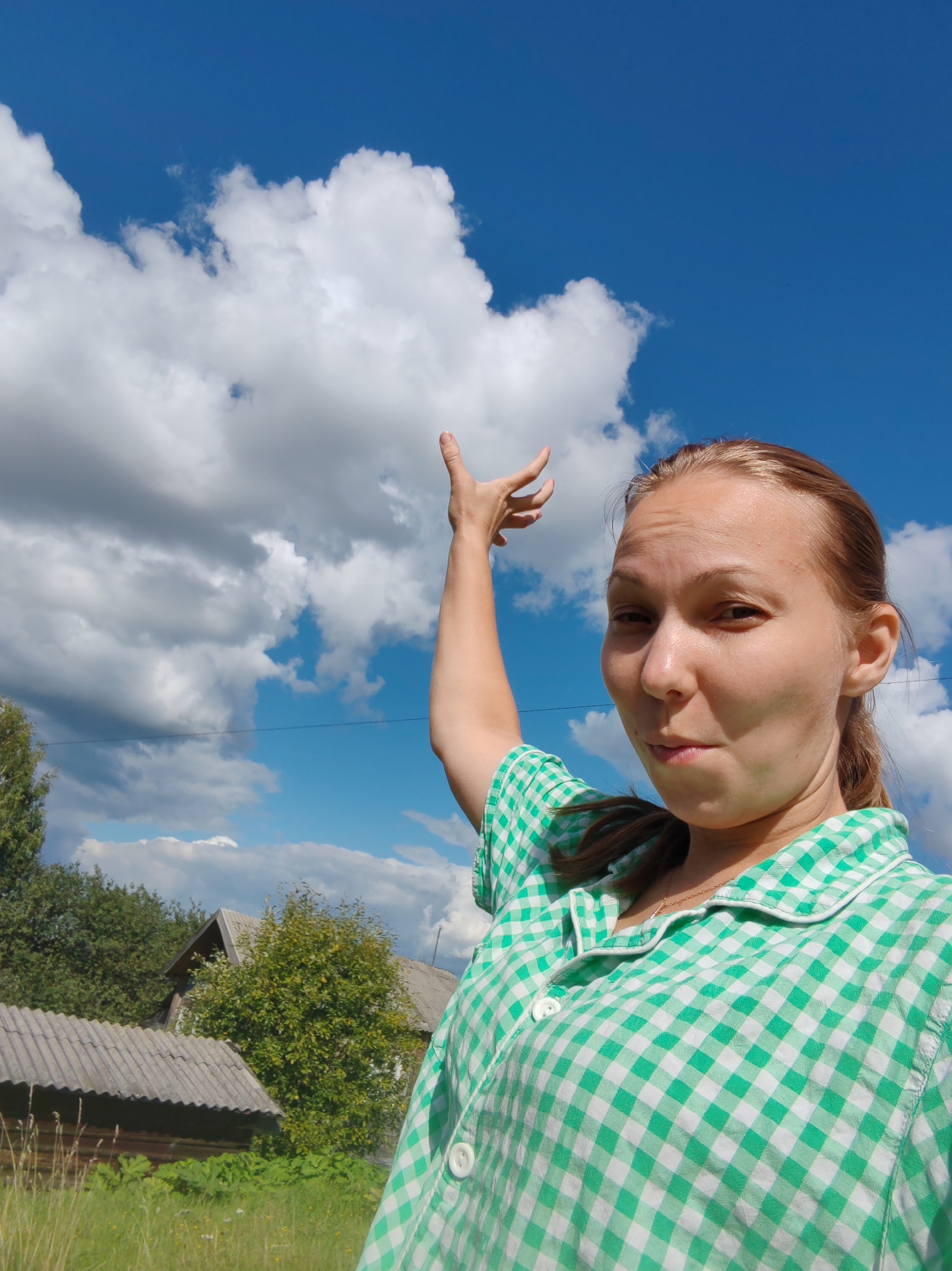 A young woman with light brown hair tied in a ponytail, wearing a green and white checkered short-sleeved shirt, is taking a selfie outdoors under a blue sky with scattered white clouds. She is positioned in the lower right quadrant of the image, with her right arm raised and pointing upwards towards a large cloud. Her facial expression is a slight pout. In the background, there are trees, two rustic wooden houses with corrugated roofs, and a grassy field. The scene appears to be a rural setting in Chukhlem, Russia.