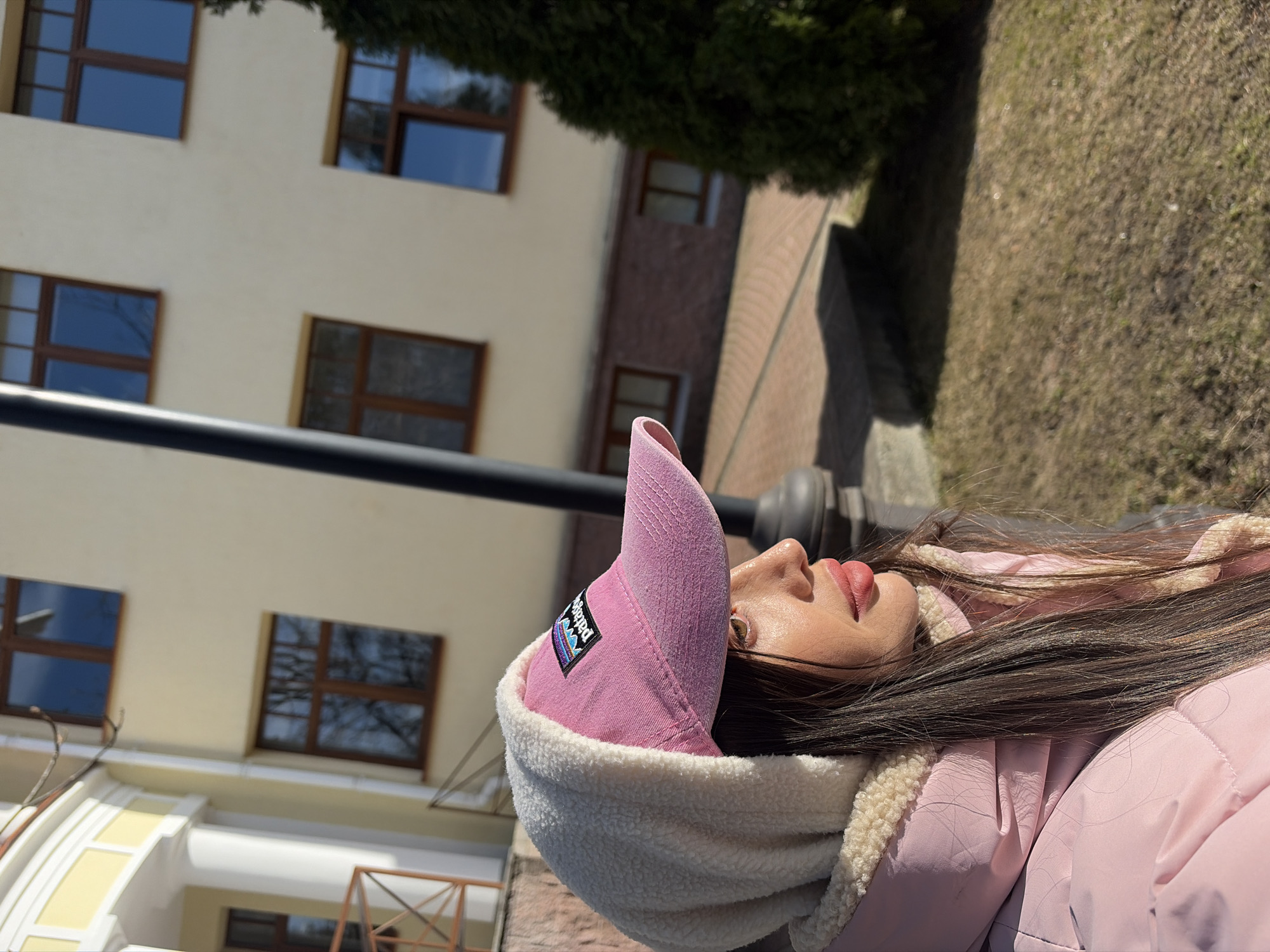 A woman with dark hair lies on a park bench in what appears to be Saint Petersburg, Russia, judging by the architecture. She wears a pink jacket and a pink baseball cap with a white fleece lining. Buildings with windows, including a brick building, are visible in the background, along with some green trees and lawn. There is a black metal object, possibly a lamp post, near the bench.