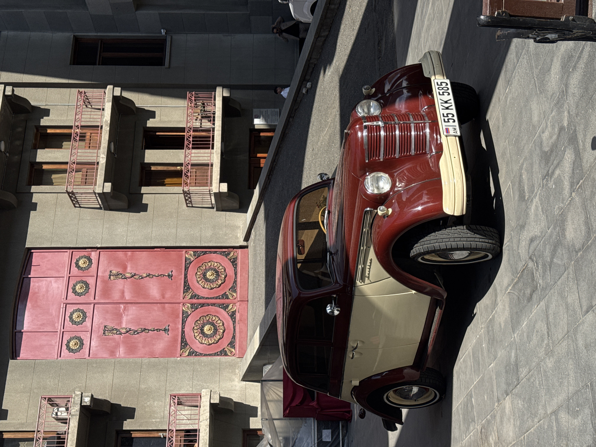 A vintage, two-toned burgundy and cream automobile is parked on a stone-tiled street bathed in bright sunlight, casting long shadows. The car, with its chrome grille and distinctive headlights, faces towards the right of the frame. A license plate with the text "AM 55 KK 585" is clearly visible on the front bumper.
Behind the car, a multi-story building with a grey facade dominates the background. The building features several balconies with ornate red railings, and large pink doors with decorative circular and floral patterns are visible on a lower level. Some windows on the building are dark, suggesting they are either closed or the interior is dimly lit.
In the upper part of the image, on the sidewalk adjacent to the street, a few individuals are standing. One person in a white shirt is looking towards the car, while another figure is partially obscured by shadow. The scene appears to be taking place outdoors during the daytime, under clear weather conditions, in what is indicated to be Yerevan, Armenia. The overall impression is one of a historical street scene, possibly a tourist attraction or a display of vintage vehicles.