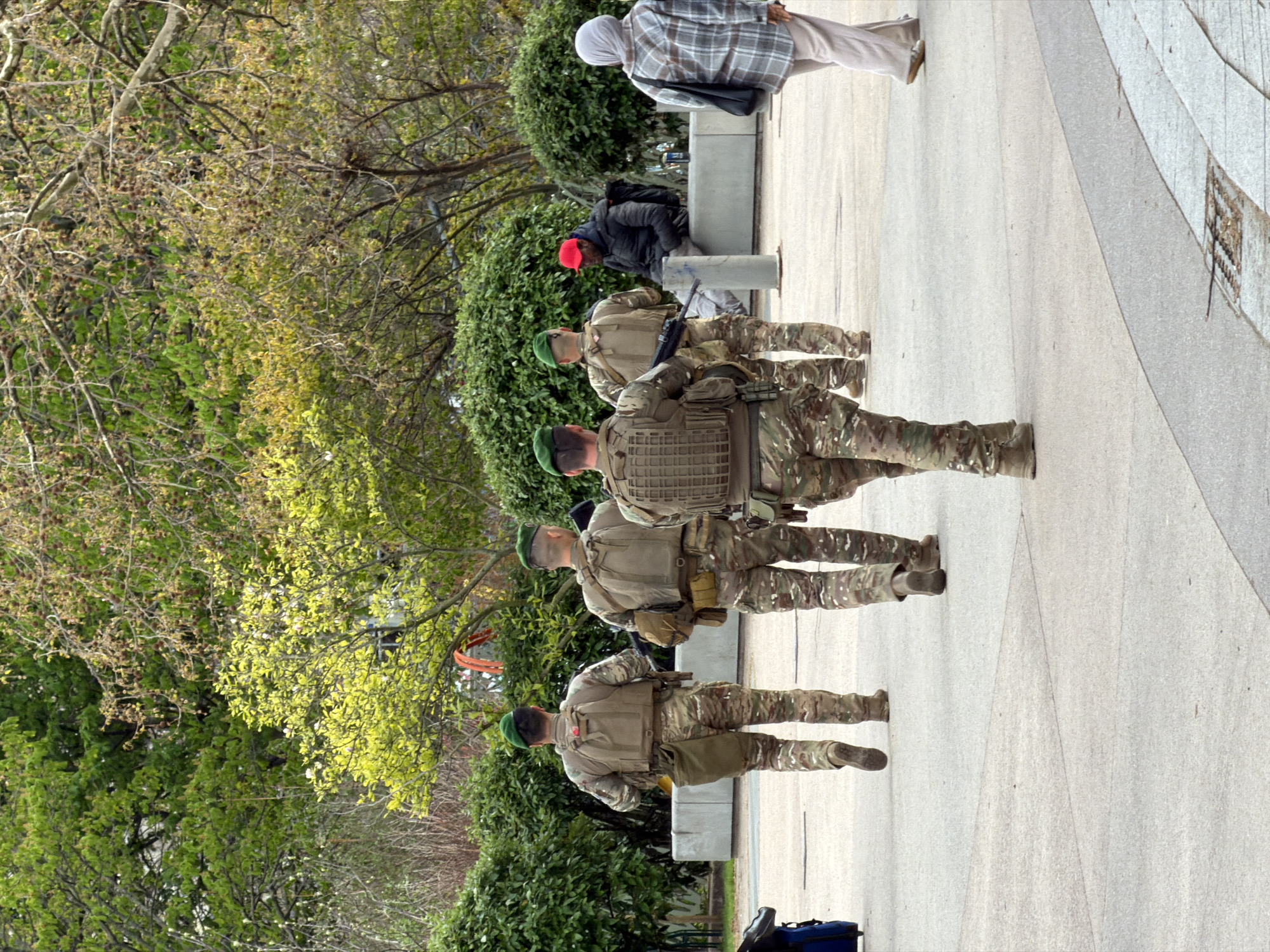 The image depicts a public urban landscape in Paris, France, characterized by wide paved walkways, manicured green bushes, and various trees, suggesting a park or large esplanade environment.
In the midground, four individuals, identified as soldiers, walk away from the camera. They are uniformly dressed in multi-camouflage fatigues, green berets, and tactical vests equipped with pouches and gear. Each soldier carries a firearm, likely an assault rifle, visible either slung across their back or held. The soldier closest to the camera has an additional large brown pouch on his left hip.
To the right, a civilian male is seated on a light grey concrete bench, wearing a dark jacket and a red baseball cap, with a black backpack behind him. He faces generally towards the left. Further to the right and slightly behind the soldiers, a civilian female walks away from the camera. She wears a light-colored headscarf, a plaid jacket, light-colored pants, and carries a dark shoulder bag.
The foreground consists of light grey concrete paving with dark linear divisions. Lush green foliage from bushes lines the pathway, and trees with varied leaf cover, some fully green, others with lighter new growth or bare branches, fill the background. A blue piece of luggage is partially visible on the ground in the far left foreground. No direct interaction between the soldiers and civilians is observed.