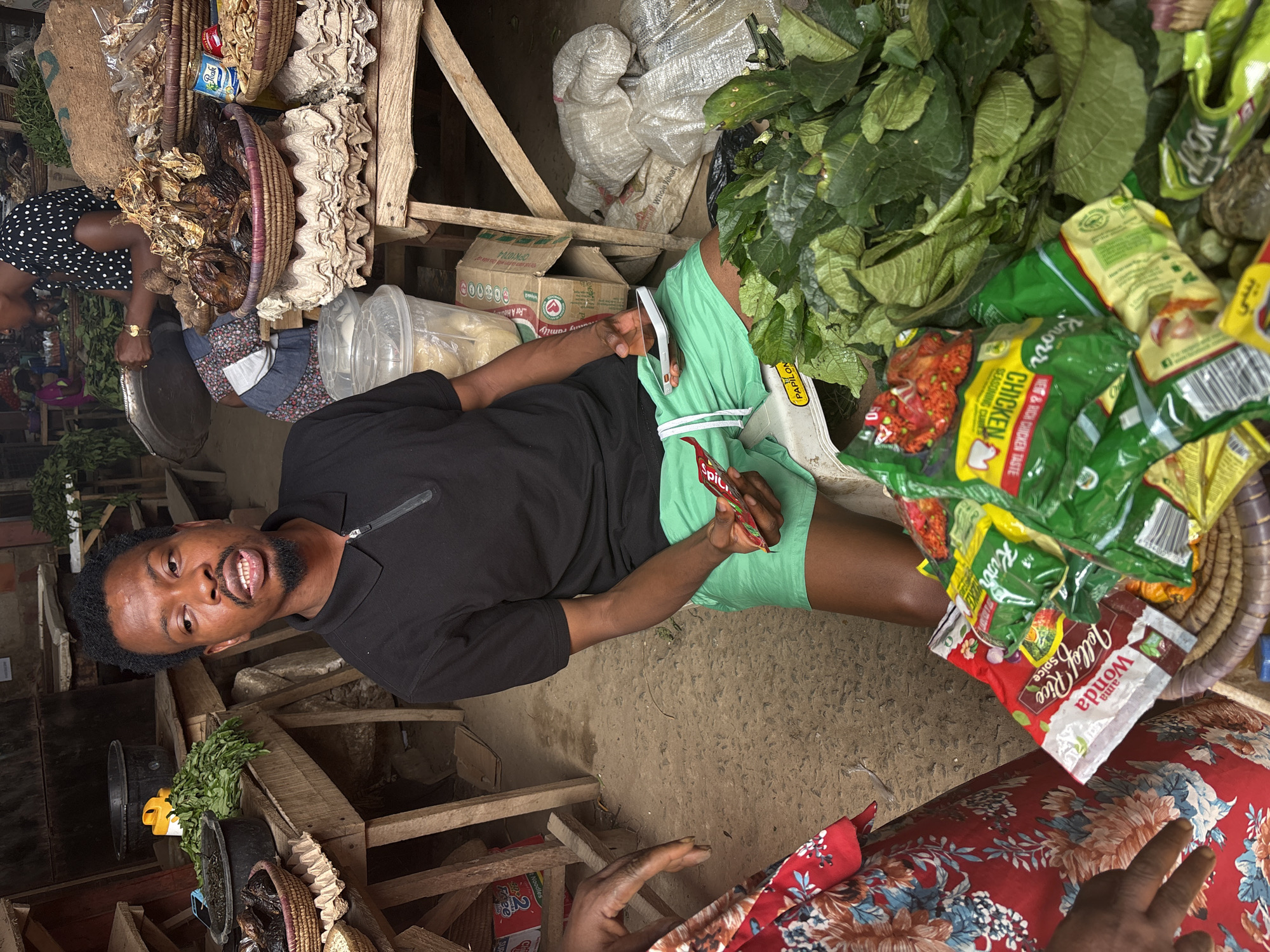 A man wearing a black polo shirt and green shorts is sitting in a market environment, holding a red Spili seasoning packet. The market scene in Iddo Tudunwada, Nigeria includes baskets containing items like dried fish and egg cartons. Packets of seasoning (Knorr, Wonda Jollof Rice) and leafy greens are also visible. The man seems to be interacting with someone wearing a red floral pattern top. Various crates and boxes, including one with "For A Healthy Family" printed on it, suggest market activities.