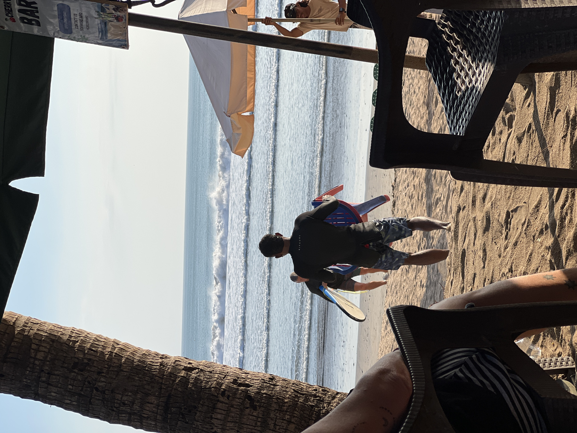 The image captures a beach scene in Kuta, Indonesia. In the foreground, two dark plastic chairs are positioned on the sand, with one occupied by a person whose legs and torso are partially visible. The other chair is empty. Behind the chairs, a sandy beach stretches towards the ocean. Three individuals are walking towards the water, two of whom are carrying surfboards. The person in the middle is wearing a black wetsuit and shorts, holding a white and blue surfboard. Another individual behind them is also carrying a surfboard. A person in a tan t-shirt and black cap is standing near a beige umbrella, holding onto its pole. The ocean is calm with gentle waves breaking on the shore. The sky above is clear and blue. A sign for "CHERRY BAR" is visible, indicating a nearby establishment. A palm tree trunk is prominent on the left side of the frame, suggesting a tropical location. The overall atmosphere is relaxed and recreational.