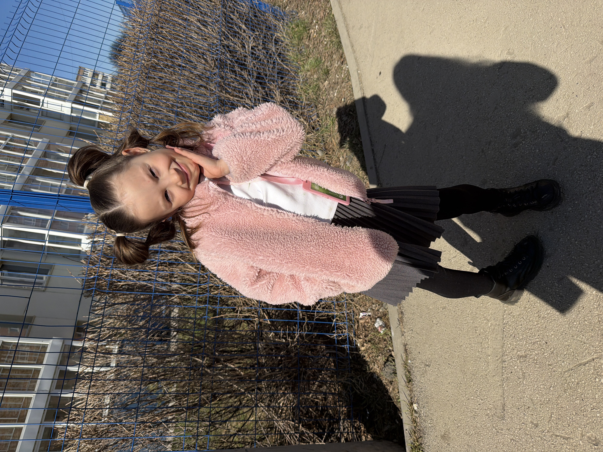 A young girl stands on a sidewalk, likely in Perm, Russia based on visible architecture. She wears a pink fuzzy jacket, a white shirt, a black pleated skirt, black tights, and black boots. Her hair is styled in pigtails. She has one hand on her face and is smiling. Behind her is a blue metal fence, a dry bush, and a building with multiple windows. The ground shows patchy grass and trash. The lighting suggests a sunny day. The shadow of a person is cast on the sidewalk.