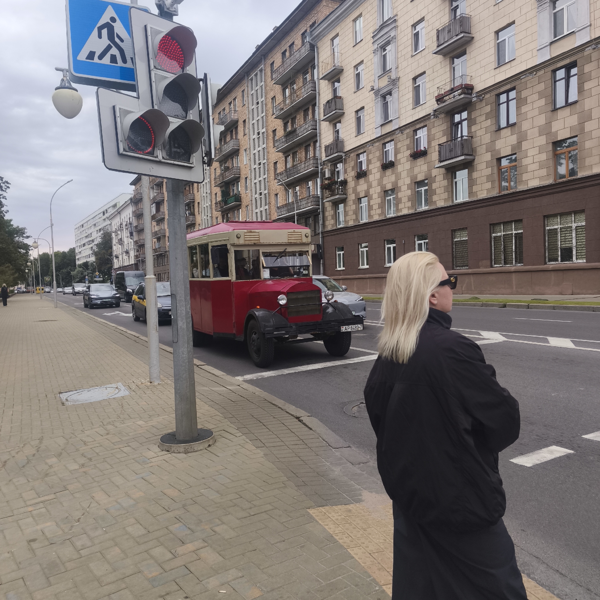 A medium shot captures a street scene in Minsk, Belarus, under an overcast sky. In the foreground, a person with blonde hair, wearing dark sunglasses and a black jacket, stands facing away from the camera, looking towards the street. The person is positioned on a brick-patterned sidewalk.
To the left, a traffic light displays a red signal for vehicles and pedestrians, accompanied by a pedestrian crossing sign. A lamppost stands on the sidewalk near the traffic light.
Dominating the mid-ground is a vintage red and beige bus with an open-top passenger section and black fenders. It is stopped at the crosswalk. Behind the bus, several cars are visible on the road, including a dark grey Tesla and a yellow taxi. The street is lined with multi-story apartment buildings with brick facades, balconies, and numerous windows. Trees are present along the sidewalk on the left side of the image. The overall environment suggests an urban setting.