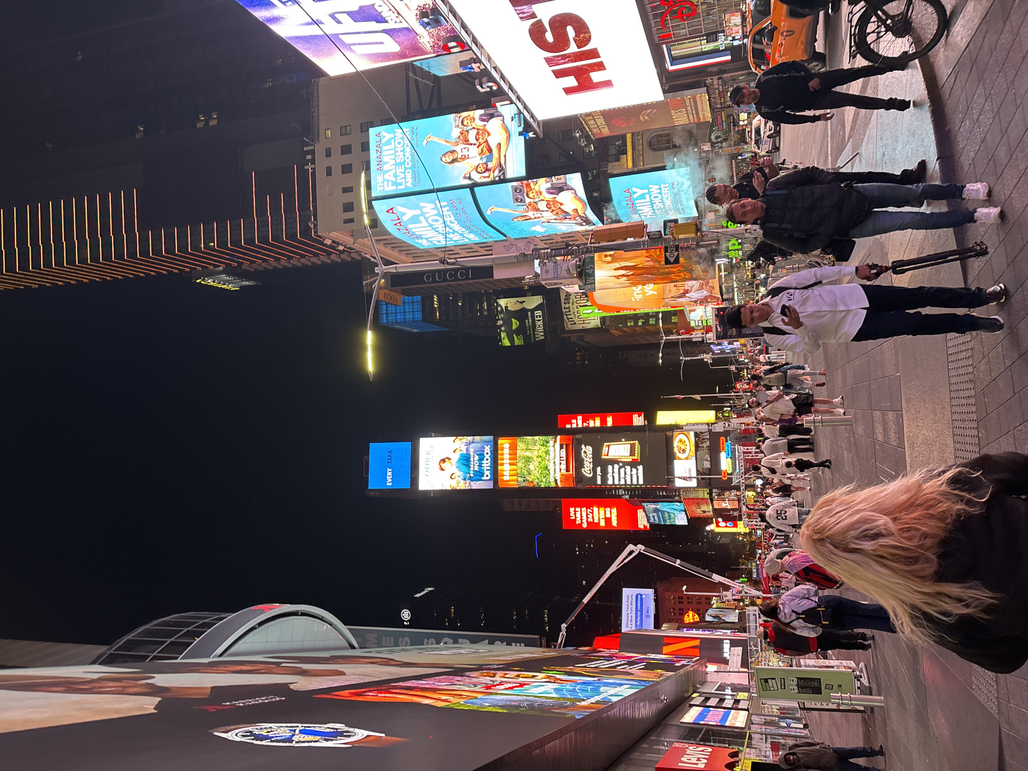 The image depicts a vibrant nighttime scene in Times Square, an iconic commercial intersection in New York City. Numerous people are present, indicating a busy urban environment. In the mid-ground, three men stand on the paved street; one wears a white shirt and holds a camera tripod, another is in a black jacket, and a third is further back near a black bicycle and a yellow taxi. In the far foreground, the back of a person with long, blonde hair is visible.
The area is dominated by an extensive array of towering, brightly lit digital billboards and screens, illuminating the surroundings against the dark night sky. These displays show various advertisements and announcements. Visible text includes "HIS," "THE ANAZALA FAMILY LIVE SHOW AND CONCERT," "GUCCI," "WICKED," "LITTLE SHOP OF HORRORS," "Coca-Cola," "britbox," "EVERY TARA," "LIVE TABLE GAMES 24/7," "MN LED," "Levis," and "Tech Debt in a New York Minute." The overall atmosphere is lively and commercial, characteristic of this famous tourist destination.
