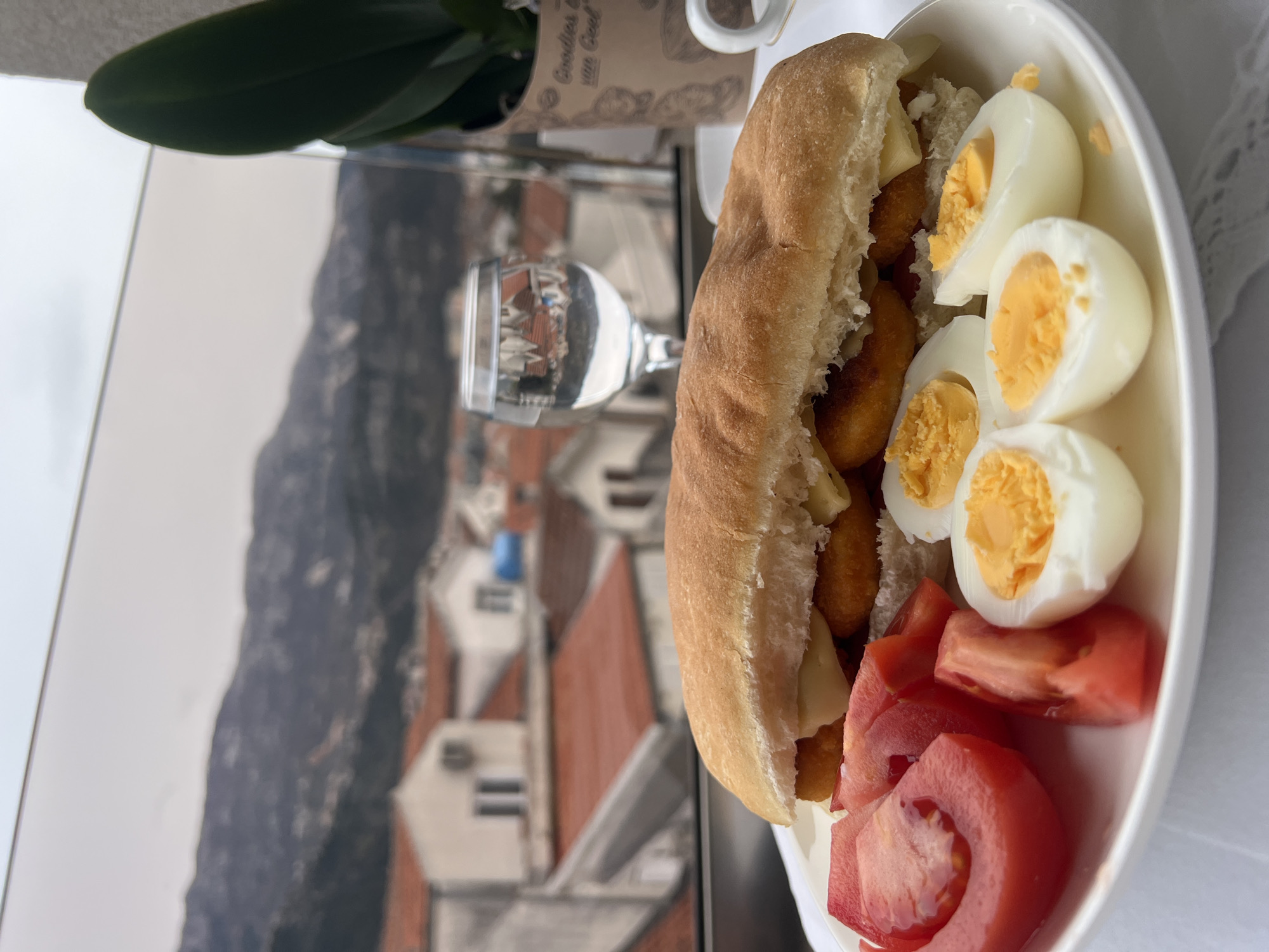 The image shows a white plate with a sandwich, hard-boiled eggs, and sliced tomatoes. The sandwich appears to contain chicken nuggets and cheese. A clear glass of liquid (likely water) stands next to the plate. In the background, outside a window, the city of Budva, Montenegro is visible, with red-tiled roofs and mountainous terrain. There is also a plant with green leaves in a decorative pot.