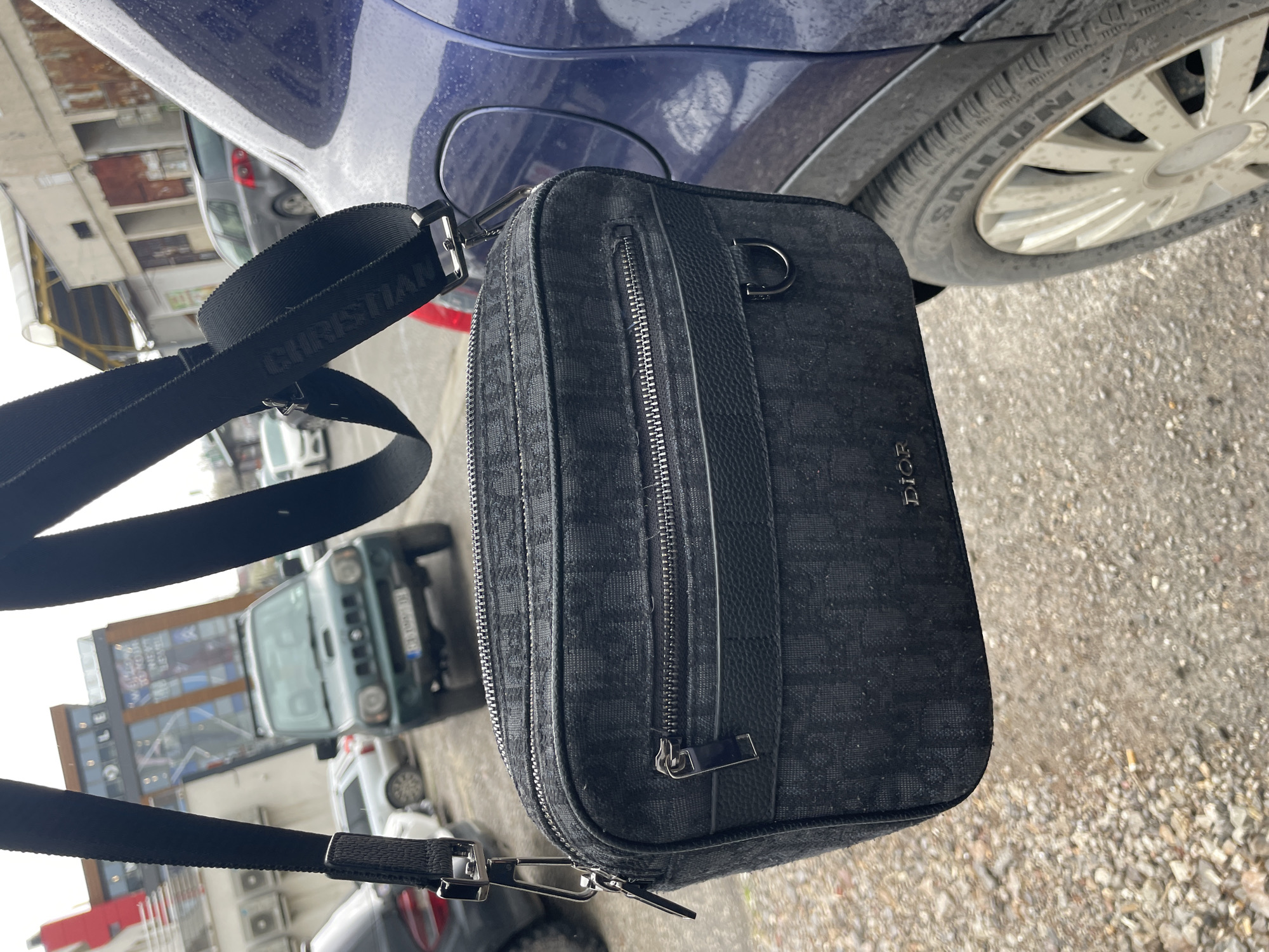 The image is a close-up shot of a black Dior messenger bag hanging from a blue car, with a "CHRISTIAN" branded strap. The background shows a street scene in what appears to be an urban setting. Key background elements include cars, buildings, and possibly a billboard. The environment appears to be a street or parking area with grey asphalt ground. The car is parked with the tire visible, and the tire has the word "SAILUN" written on it. There are no people directly visible in the frame. The location is Sofia, Bulgaria.