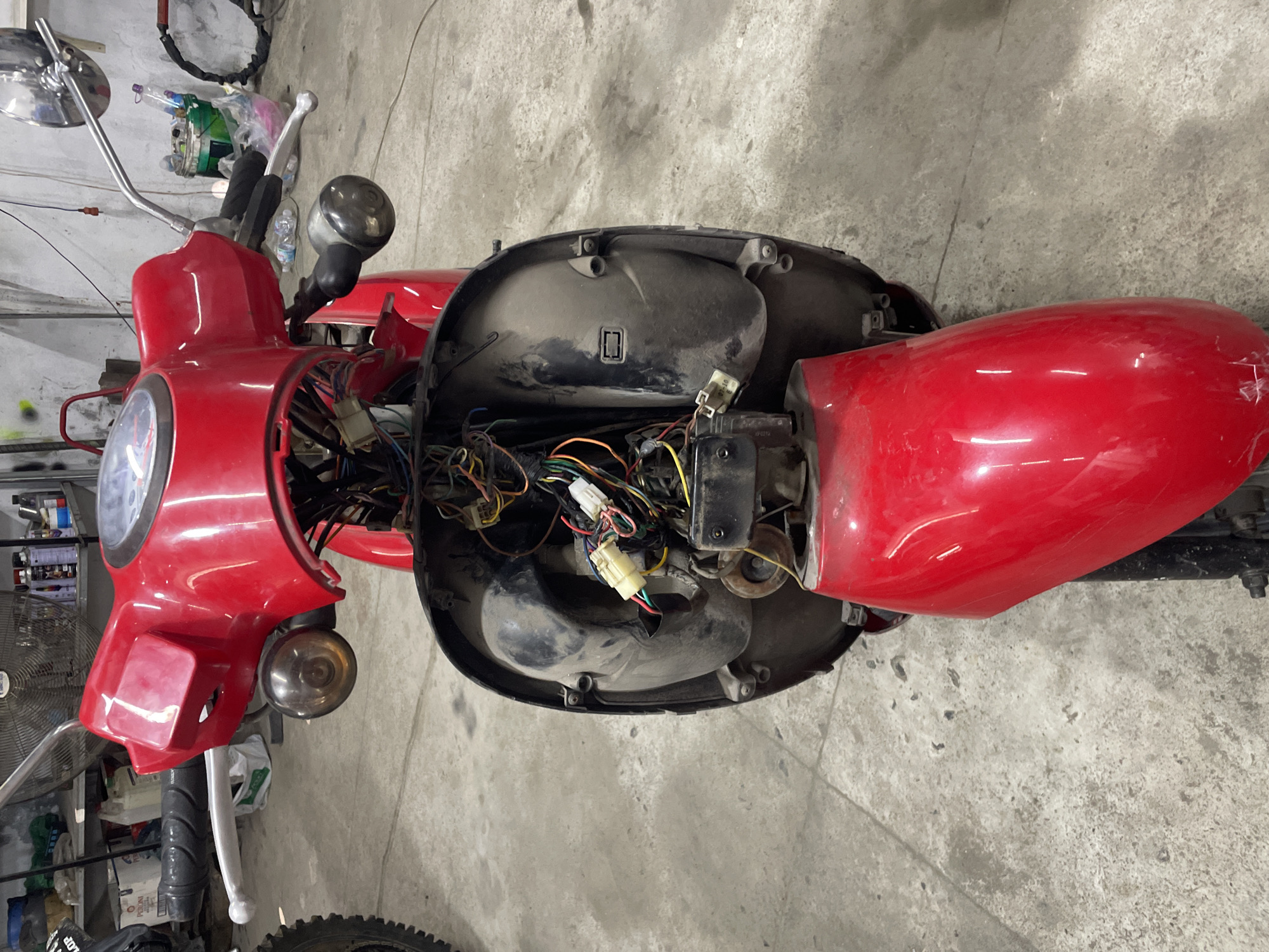 The image shows a red scooter undergoing repairs in a garage or workshop setting. The scooter's front body panels are removed, exposing the wiring harness and other internal components. The surrounding environment is a concrete floor with evidence of tools and spare parts nearby. The location in Bac Nha Trang Ward, Vietnam is given, but there are no visual indicators to confirm this location within the image.