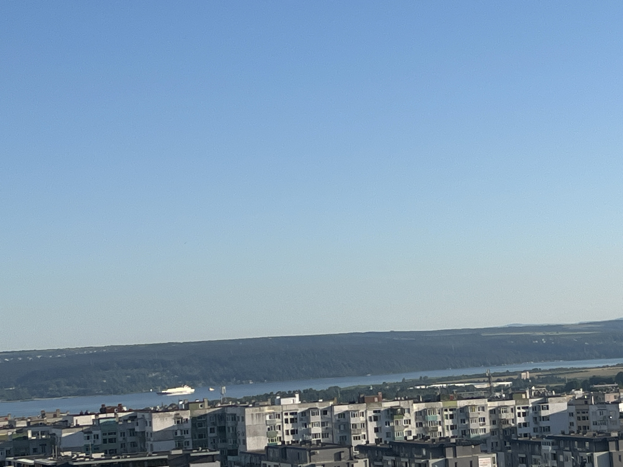 This is a high-angle, wide shot of the city of Varna, Bulgaria, on a clear, bright day. The image captures a panoramic view of the city's skyline, dominated by a vast expanse of apartment buildings in the foreground. These buildings are characterized by their utilitarian, Soviet-era architecture, with repetitive window patterns and varied color palettes, including white, grey, and green accents.
Beyond the city, a wide, tranquil river or bay stretches out, reflecting the clear blue sky. A lone white ferry or cargo ship is visible in the middle distance, traversing the water. The opposite bank of the river is lined with a dense, dark green forest that slopes upwards to meet a hazy, undulating horizon, suggesting rolling hills or distant mountains. The sky above is a consistent, pale blue, with no clouds visible, indicating good weather conditions. The overall impression is one of a coastal urban environment meeting a natural landscape under a serene sky.