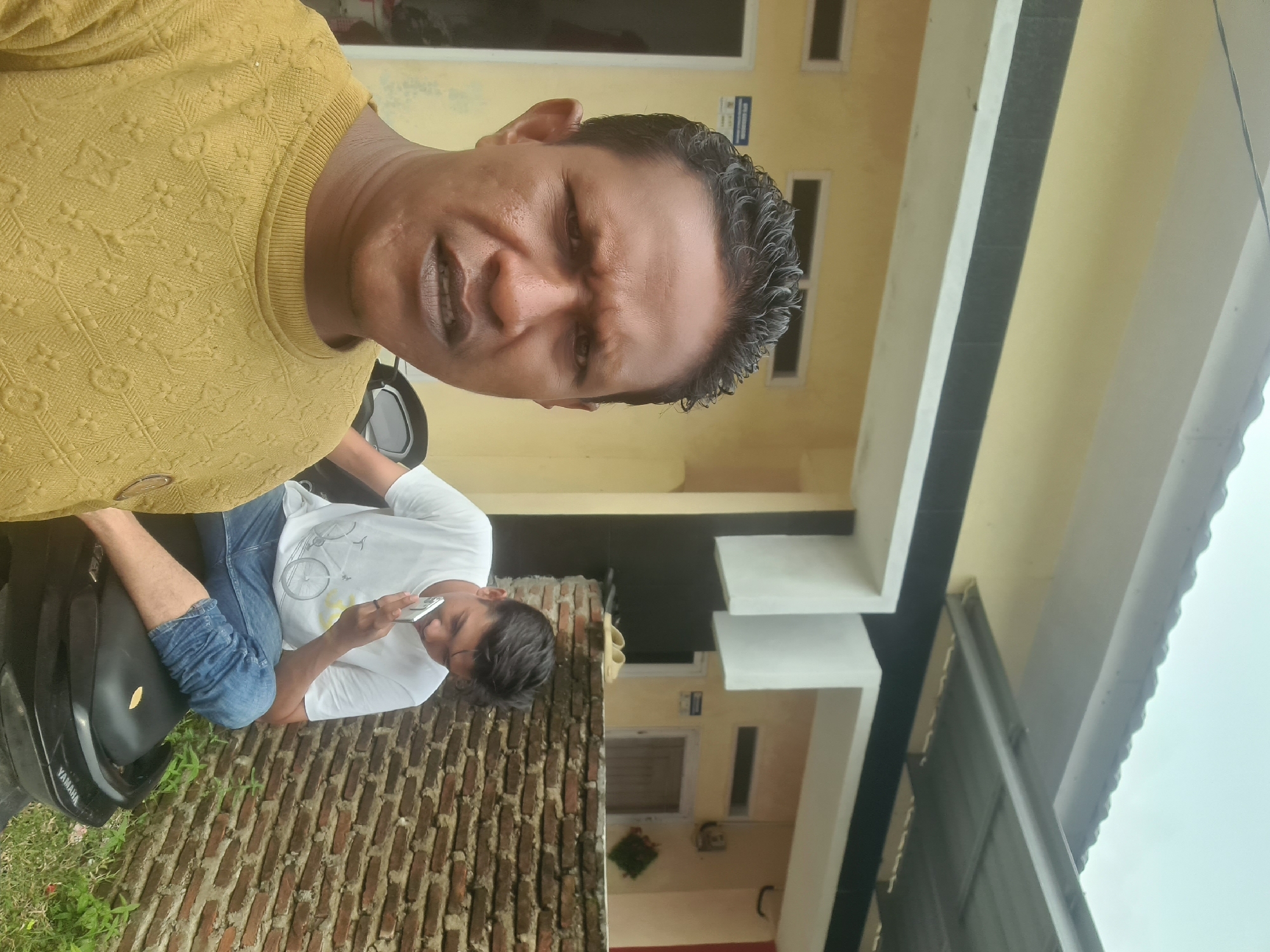 Two men are in front of a building in Lhokseumawe, Indonesia. The man in the foreground is facing the camera and wearing a yellow long-sleeved shirt with a pattern. He has dark, curly hair and is looking directly at the camera with a serious expression. Behind him, the second man is sitting on a black motorcycle. He is wearing a white t-shirt with a bicycle graphic and blue jeans. He is holding a silver smartphone to his ear, appearing to be on a call. The background shows a light yellow building with a black stripe and a white overhang. A brick wall is visible to the right of the building. Green plants grow in front of the brick wall. The sky is overcast.
