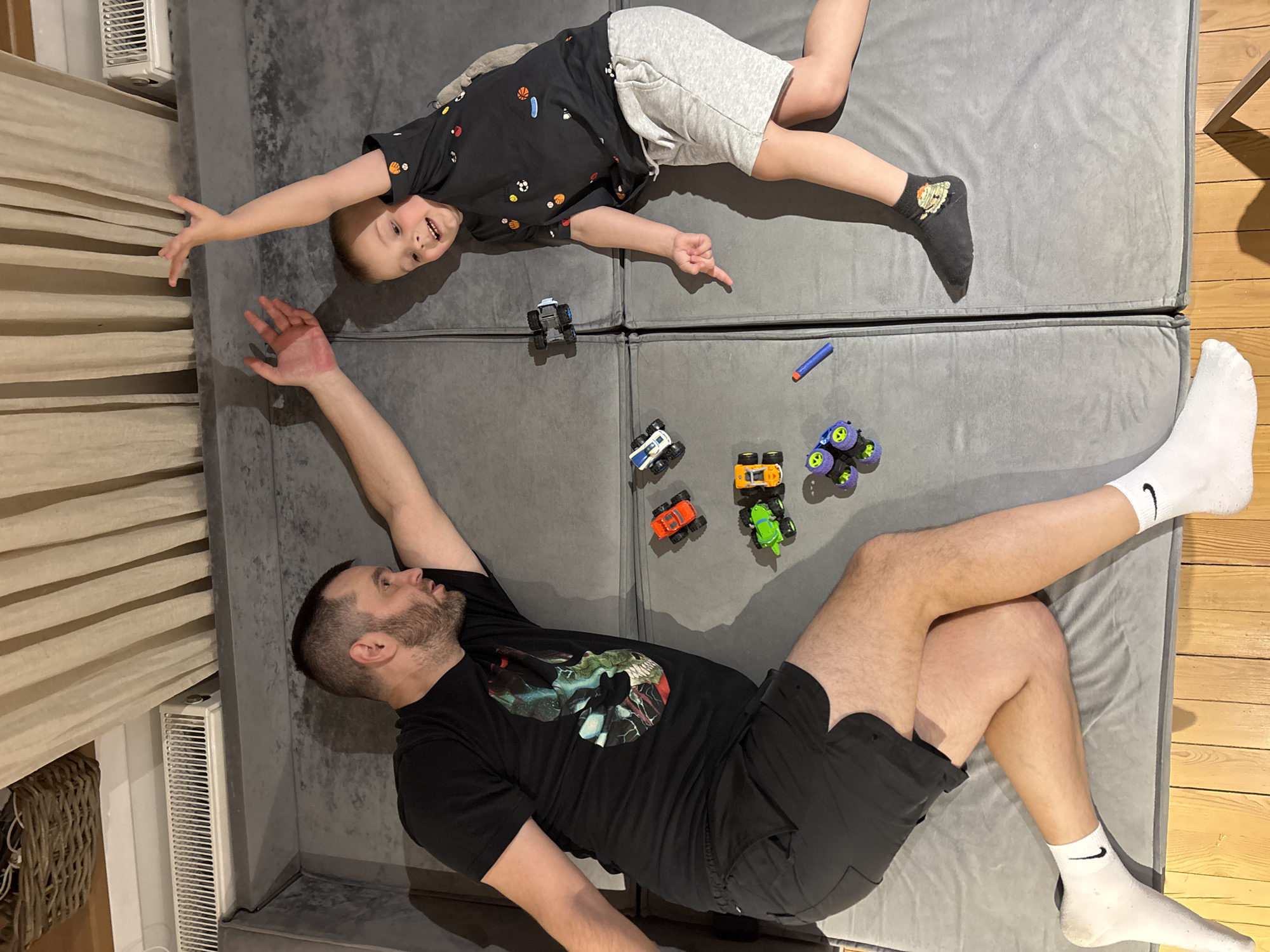 A top-down shot depicts a man and a young boy lying on a gray, cushioned surface. The man, wearing a black t-shirt with a design and black shorts, is positioned at the bottom of the frame with his head near the bottom left corner. He has a beard and is wearing white socks with a black Nike logo. His left arm is extended upwards. A young boy is lying towards the top of the frame, wearing a black t-shirt with various sports ball designs, gray shorts, and one black sock with an image. His right arm is extended, and he is looking towards the camera.
Between them, there are several toy monster trucks, including blue, orange, green, and purple ones, as well as a blue dart. A radiator is visible in the top left corner of the frame. The surface they are lying on appears to be a modular sofa or interconnected cushions.
The location is Chaikovsky, Russia.