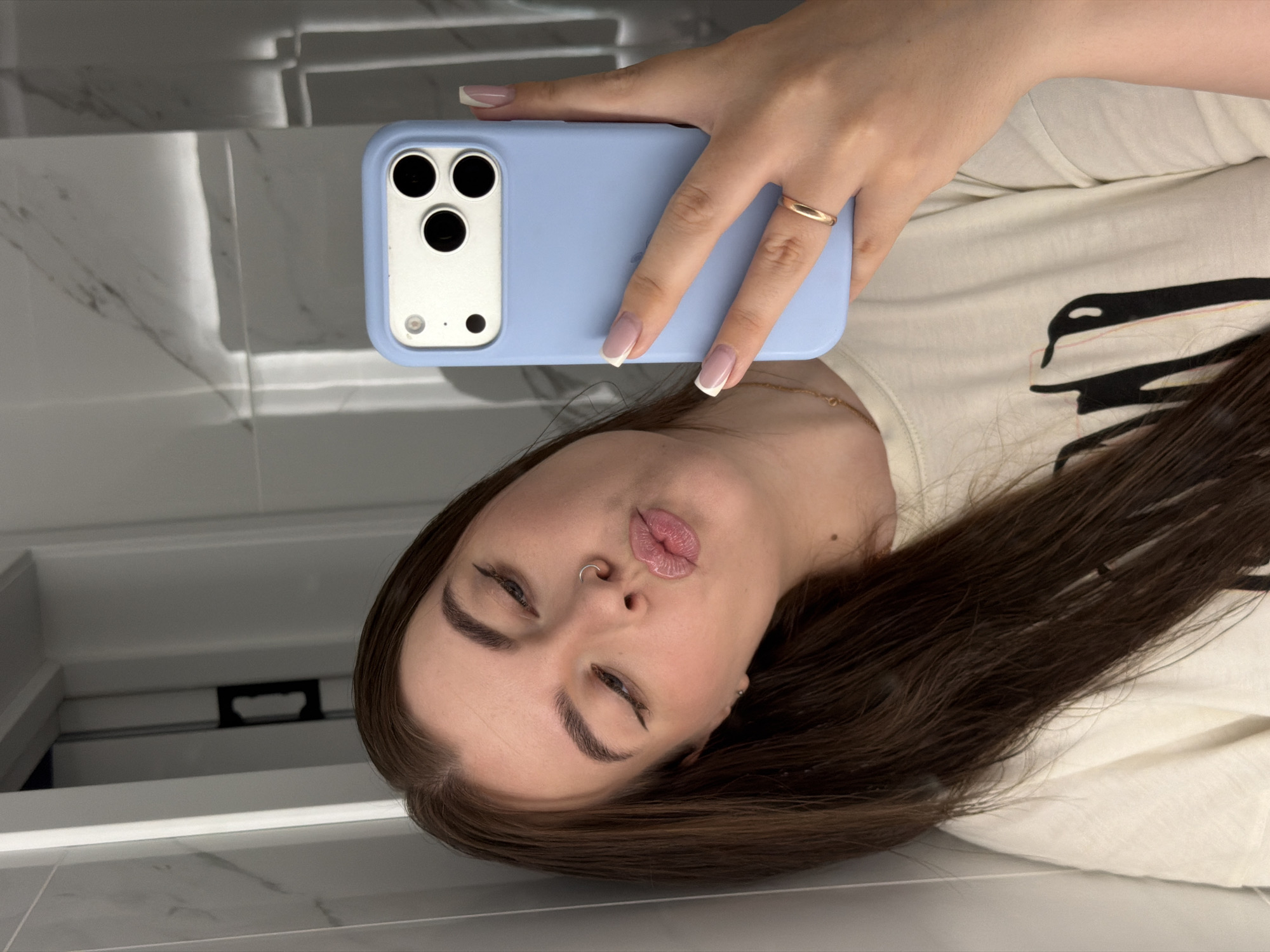A young woman with long brown hair and light makeup is taking a selfie in a brightly lit bathroom. She is wearing a light-colored t-shirt and has a nose ring and a gold ring on her finger. Her phone, with a light blue case, is held up to take the photo. The background shows tiled walls and a doorway. The image is undated, but it is likely from the city of Chaikovsky, Russia.