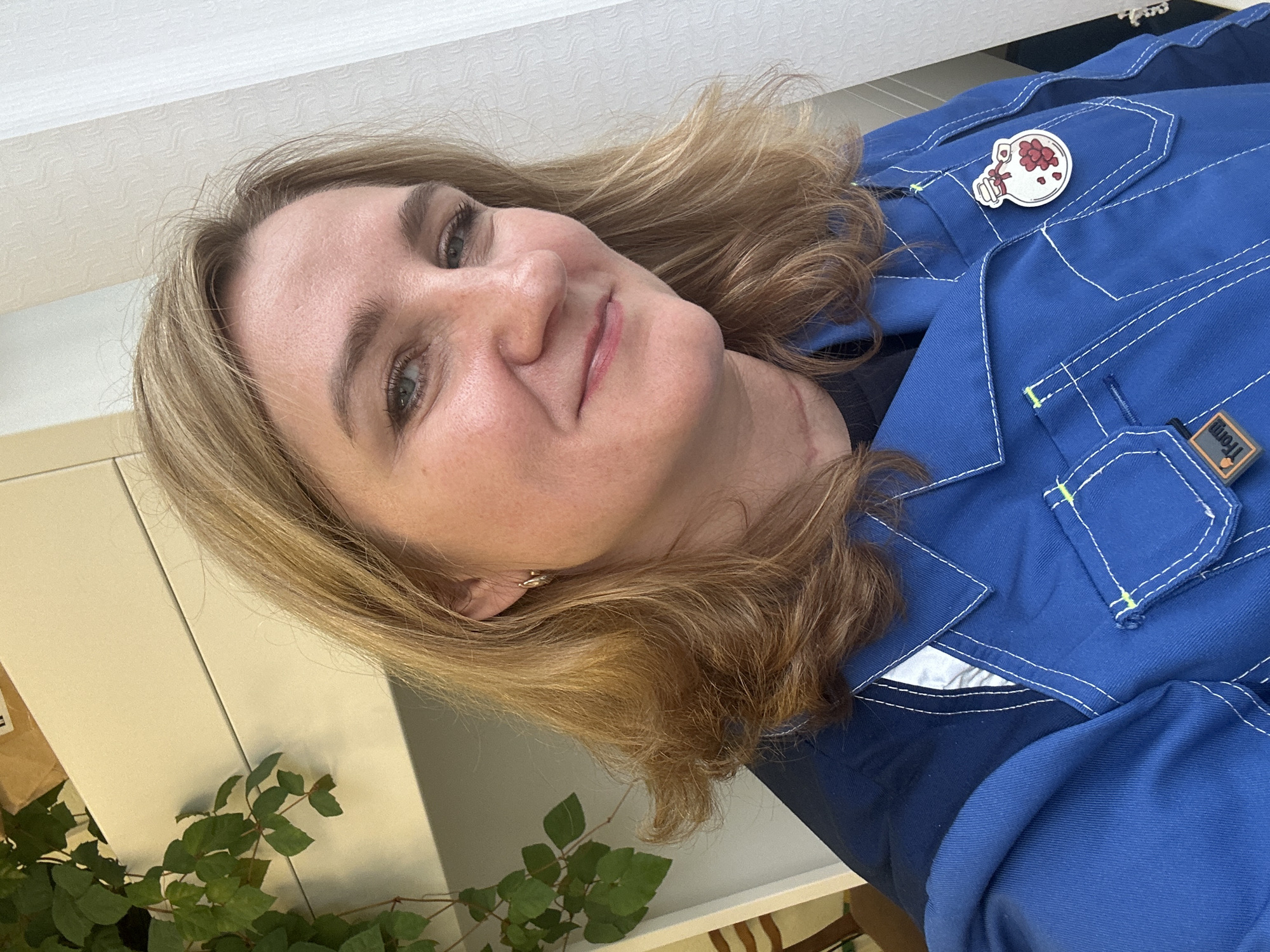 A fair-skinned woman with light brown, shoulder-length hair is posing for a selfie. She is wearing a blue jumpsuit with white stitching and a white pin depicting a brain with flowers. The photo appears to be taken indoors, near a white cabinet with a potted plant in the lower left corner. The location is Chaikovsky, Russia.