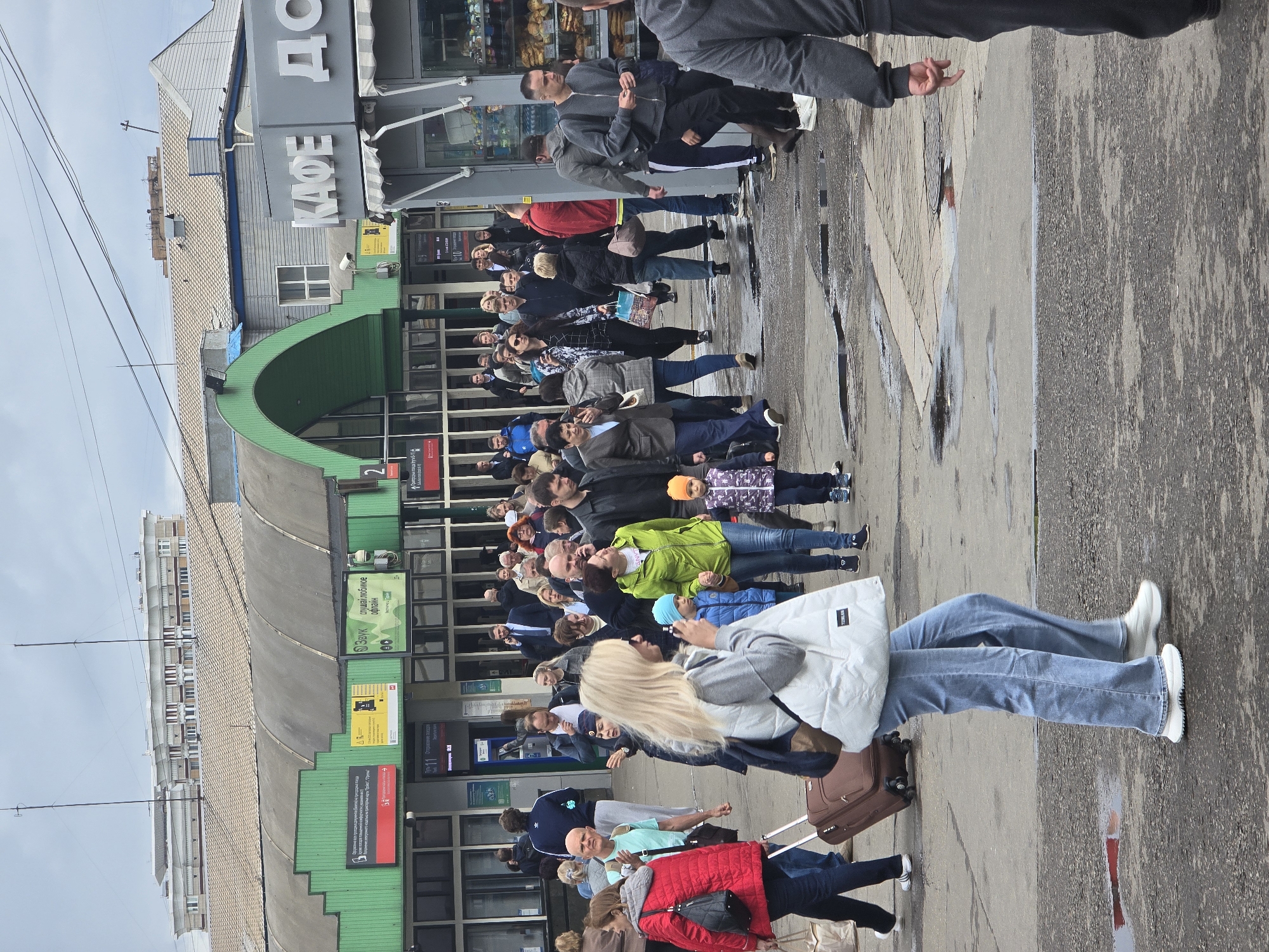 A crowd of people is lined up outside a building with a green awning and gray walls in Moscow, Russia. The sign above the entrance of the building has Cyrillic text, one word appears to read "KAFE". A bakery and restaurant are located to the right of the building. The people waiting are diverse in age and attire. Most are standing, but a few are walking. Some have luggage. The ground appears wet. The weather appears overcast.