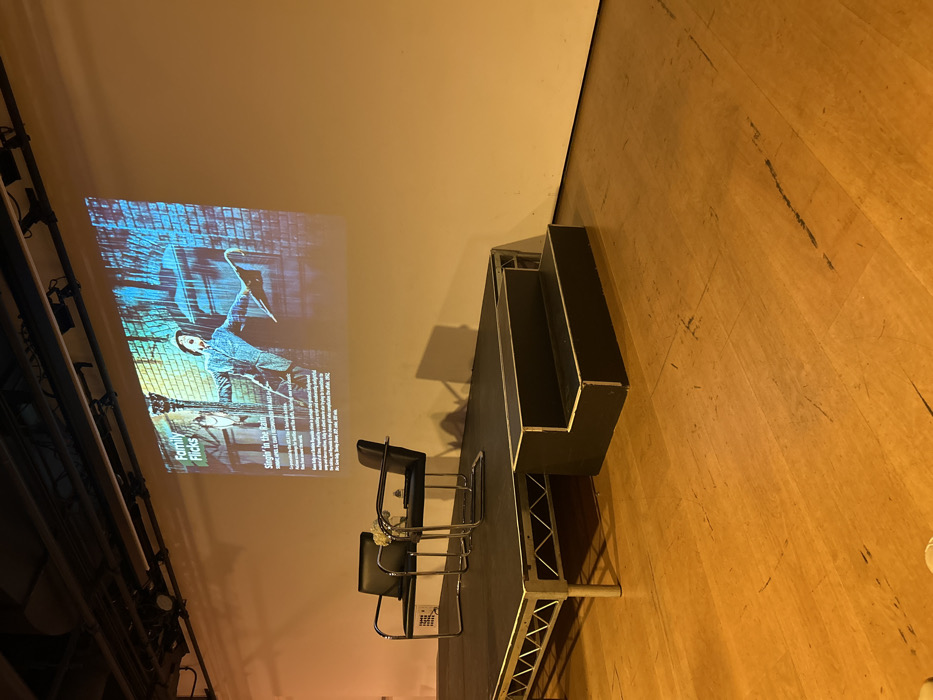 A projected image on a light-colored wall displays the title "Family Flicks" and text related to "Singin' in the Rain." Adjacent to the wall is a raised platform or stage with two black chairs and a black tiered structure. The platform's legs are exposed, revealing a metal framework. The floor is wooden, and lighting fixtures are visible along the wall's edge.