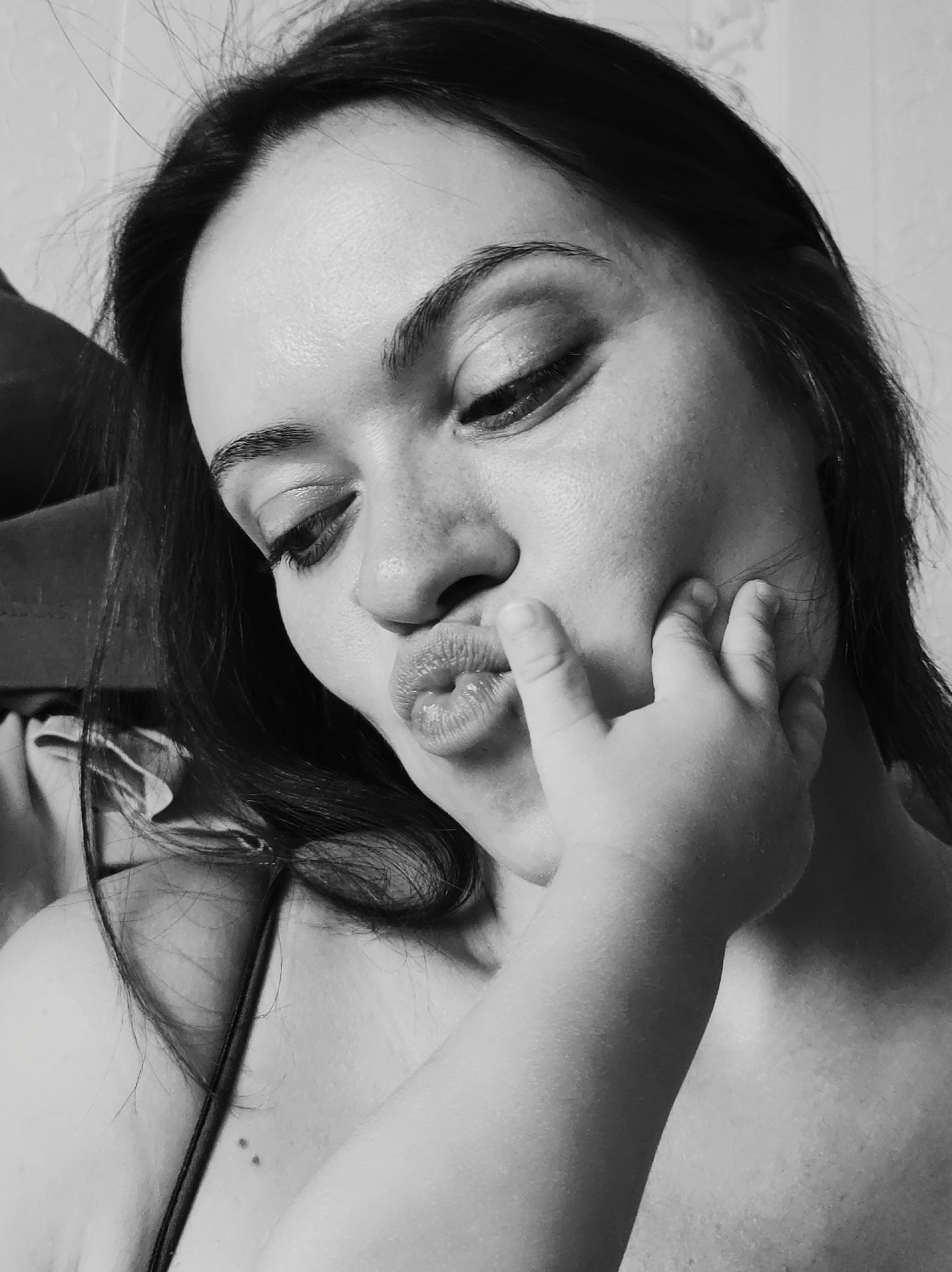 This is a black and white close-up selfie of a young woman. She is looking down with her eyes closed and her lips puckered, as if blowing a kiss. A small hand is touching her cheek, suggesting the presence of a child. The background is a plain, lightly textured wall. The lighting is soft, creating a gentle mood. The image is likely taken indoors, given the lack of natural light cues. The woman's expression conveys a sense of affection and playfulness. There is no visible text in the image. The location context provided is Chaikovsky, Russia.