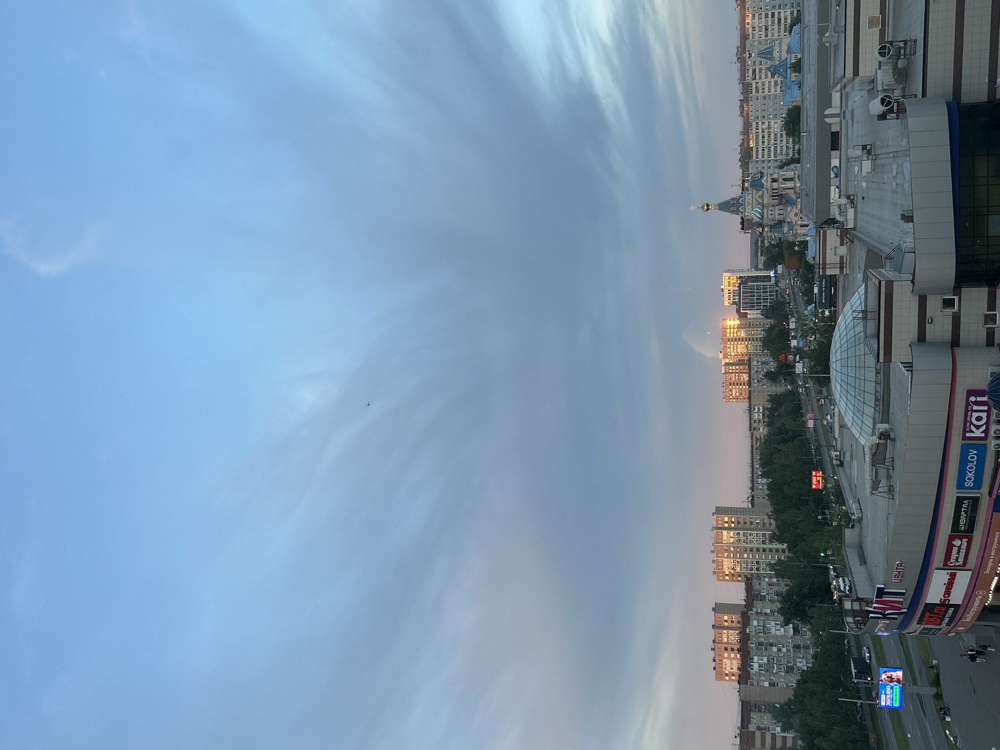 Here is a description of the image:
The image presents an elevated perspective of a cityscape in Izhevsk, Russia, under a sky transitioning from light blue to shades of white. The horizon is marked by the tops of residential and commercial buildings, including a cathedral. The foreground contains the edge of a building featuring commercial signage including brands like Sokolov and Kari. A blue electronic billboard is visible along a road, partially obscured. The time appears to be dusk or dawn, with soft light illuminating the buildings. There are hints of green foliage between the buildings, suggesting urban greenery.