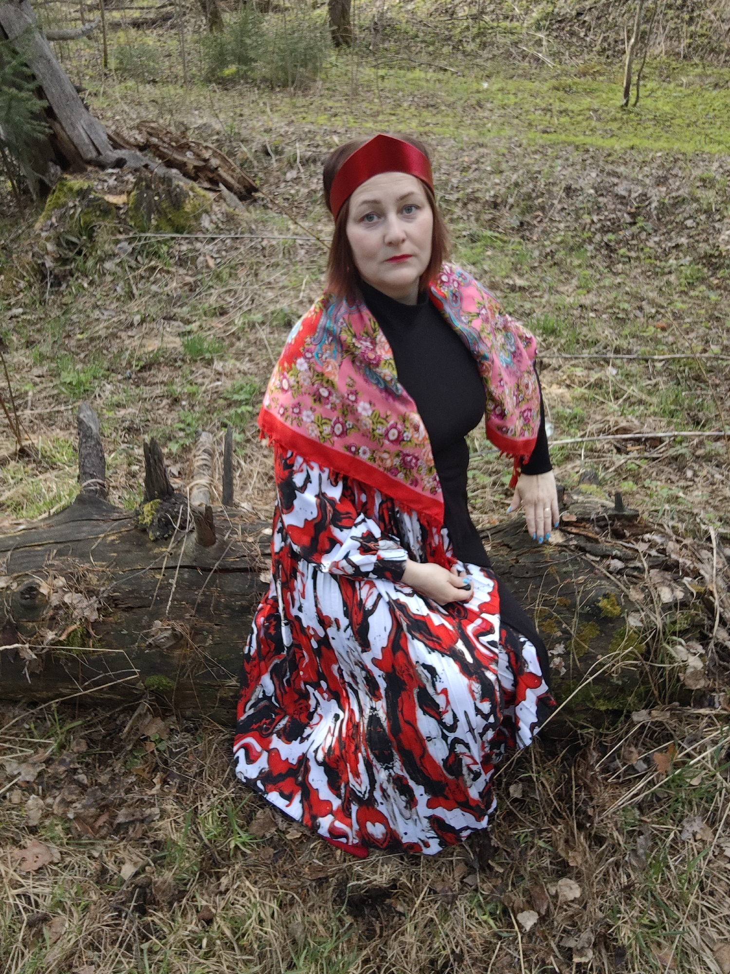 A woman is sitting on a fallen tree trunk in a wooded area in Марковский, Russia. She wears a black long-sleeved dress, a red headband, and a large scarf with floral patterns draped over her shoulders. Her skirt is a vibrant mix of red, white, and black. The ground is covered in dry leaves and grass. The background shows more trees, completing the forest setting.