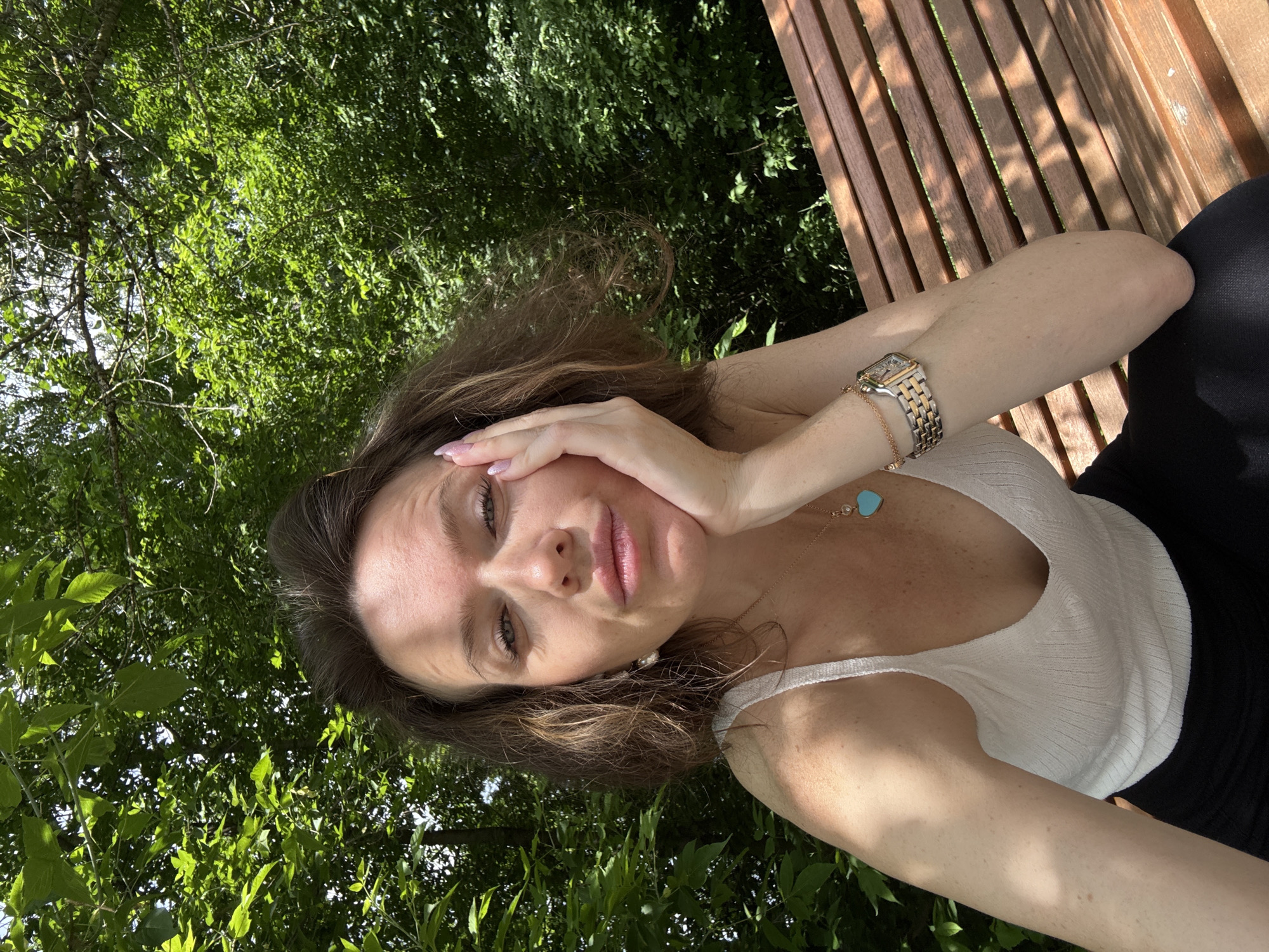 A selfie-style portrait captures a woman outdoors amidst lush greenery on a sunny day. She has medium-length brown hair, light eyes, and fair skin, looking directly at the camera with a relaxed or contemplative expression. Her right hand gently supports her head, showcasing light pink manicured nails. She wears a white ribbed tank top, a gold-toned watch with a rectangular face and metal band on her left wrist, and a delicate gold chain necklace with a light blue heart-shaped pendant.
The background is dominated by vibrant green foliage from trees, dappled with sunlight, indicating a bright day. To her right, a wooden slatted bench is visible, and a glimpse of black fabric, likely her attire, appears in the lower right. The scene suggests a tranquil moment in a park or garden, consistent with the provided location context of Moscow, Russia. There is no visible text in the image.