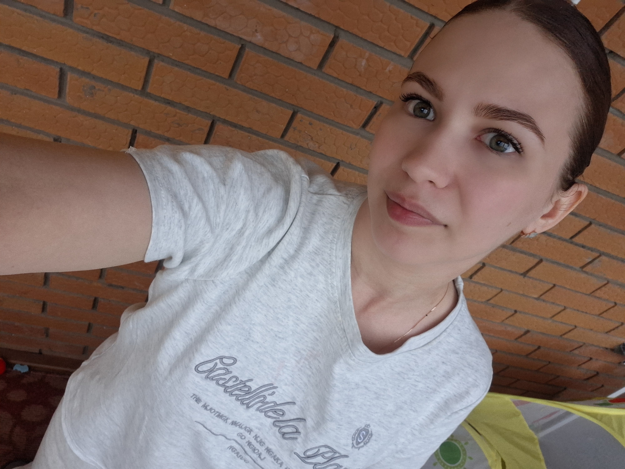 A selfie shows a fair-skinned woman with brown hair pulled back, wearing a light gray t-shirt with a faded logo on the left chest. She has light brown eyebrows, green eyes, and minimal makeup. The background is a brick wall, indicating an outdoor setting. Based on the available information, the image cannot be placed in the city of Chaikovsky, Russia.