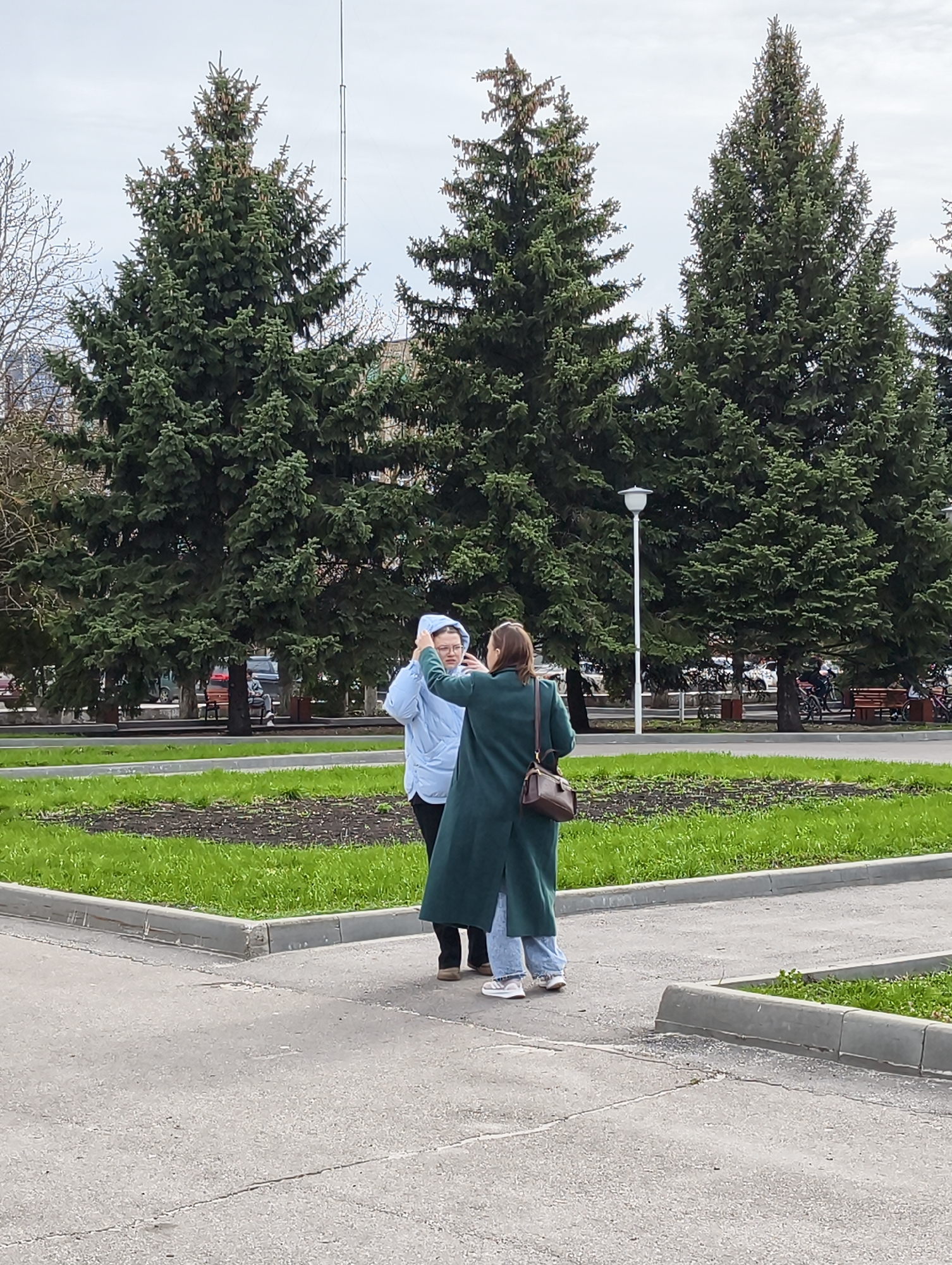 Two women stand on a paved surface in what appears to be a park or public square. The woman on the left wears a light blue hooded jacket and black pants; she is touching her hair or glasses with her right hand. The woman on the right wears a long, dark green coat over light blue jeans and carries a brown shoulder bag. They appear to be interacting with each other. Behind them are three large evergreen trees. A park bench, street lamp, and what appears to be a building are visible in the background. The sky is overcast.
Based on the metadata and analysis of street-level imagery and public landmarks in relation to the provided visual information, this image is very likely to have been captured in the vicinity of the park in the city of Balashov, Russia, considering the architecture, tree varieties, street furniture and park designs prevalent in the region. However, lacking finer details or landmarks visible in the original image, a definitive pinpoint location is not possible.