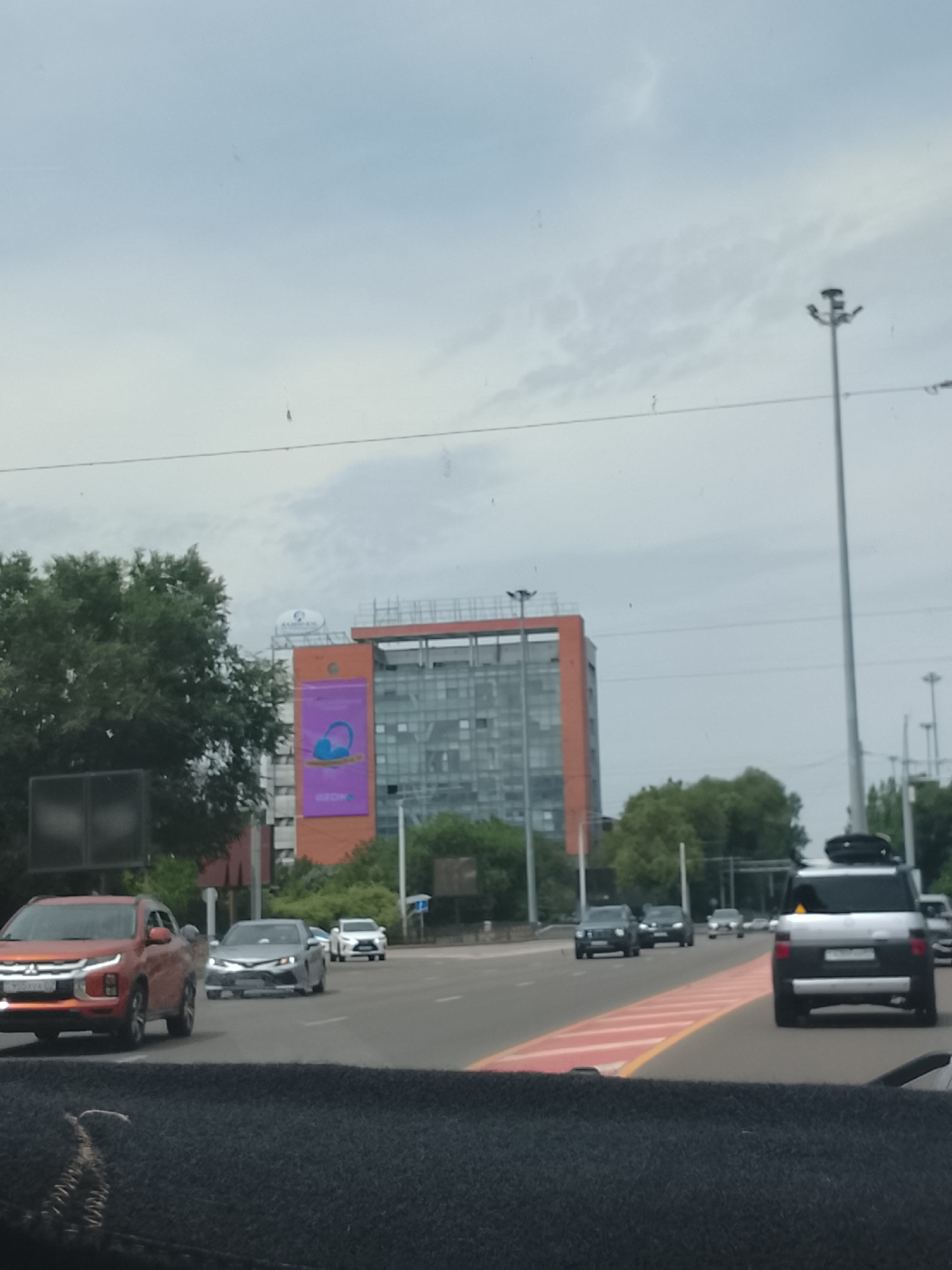 The image depicts a street scene in Almaty, Kazakhstan, under an overcast sky. A multi-story orange and glass building is prominent, featuring a large purple banner with a graphic of headphones and text. Several cars are visible on the road, including a red SUV in the foreground, a silver sedan, and a dark SUV. Trees line the sides of the road, and a tall street lamp stands on the right. The perspective suggests the photo was taken from inside another vehicle.