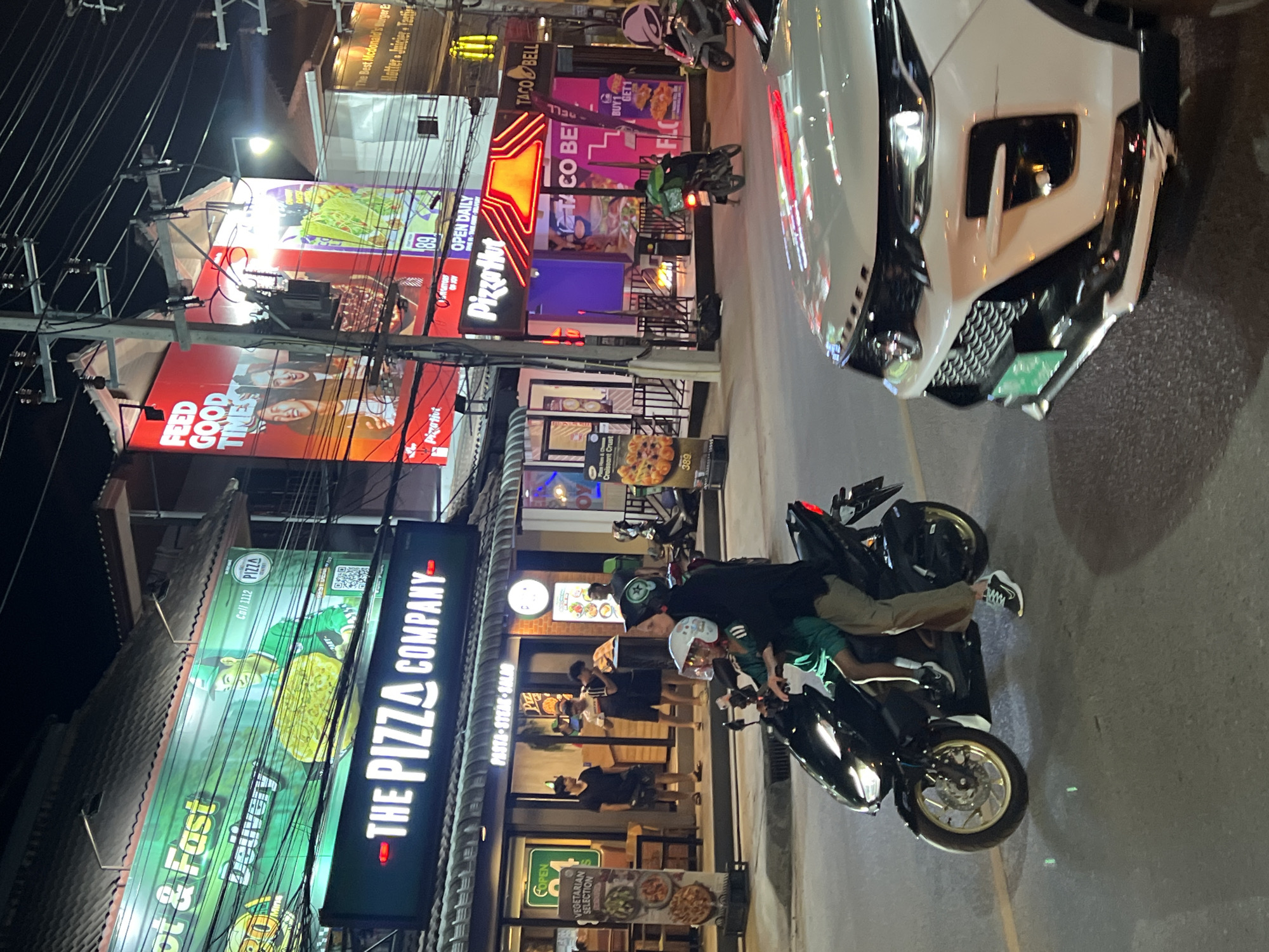 The image shows a street scene at night in Baan Bo Phut, Thailand, with several fast-food establishments visible. In the foreground, two people are riding on a black scooter. The person in front is wearing a white helmet and a green outfit, while the person behind them is wearing a black top and khaki pants, also with a helmet. To the right, the front of a white car is visible.
The background features illuminated signage for various restaurants. Prominently displayed are signs for "The Pizza Company," "Pizza Hut," and "Taco Bell." A large red billboard for Pizza Hut with the text "FEED GOOD TIMES" is also visible. Other signs suggest additional food offerings, including pizza and vegetarian selections. People are seen standing outside The Pizza Company. The street is paved, and utility poles with numerous wires run overhead. The overall environment suggests a commercial area with active businesses during nighttime hours.