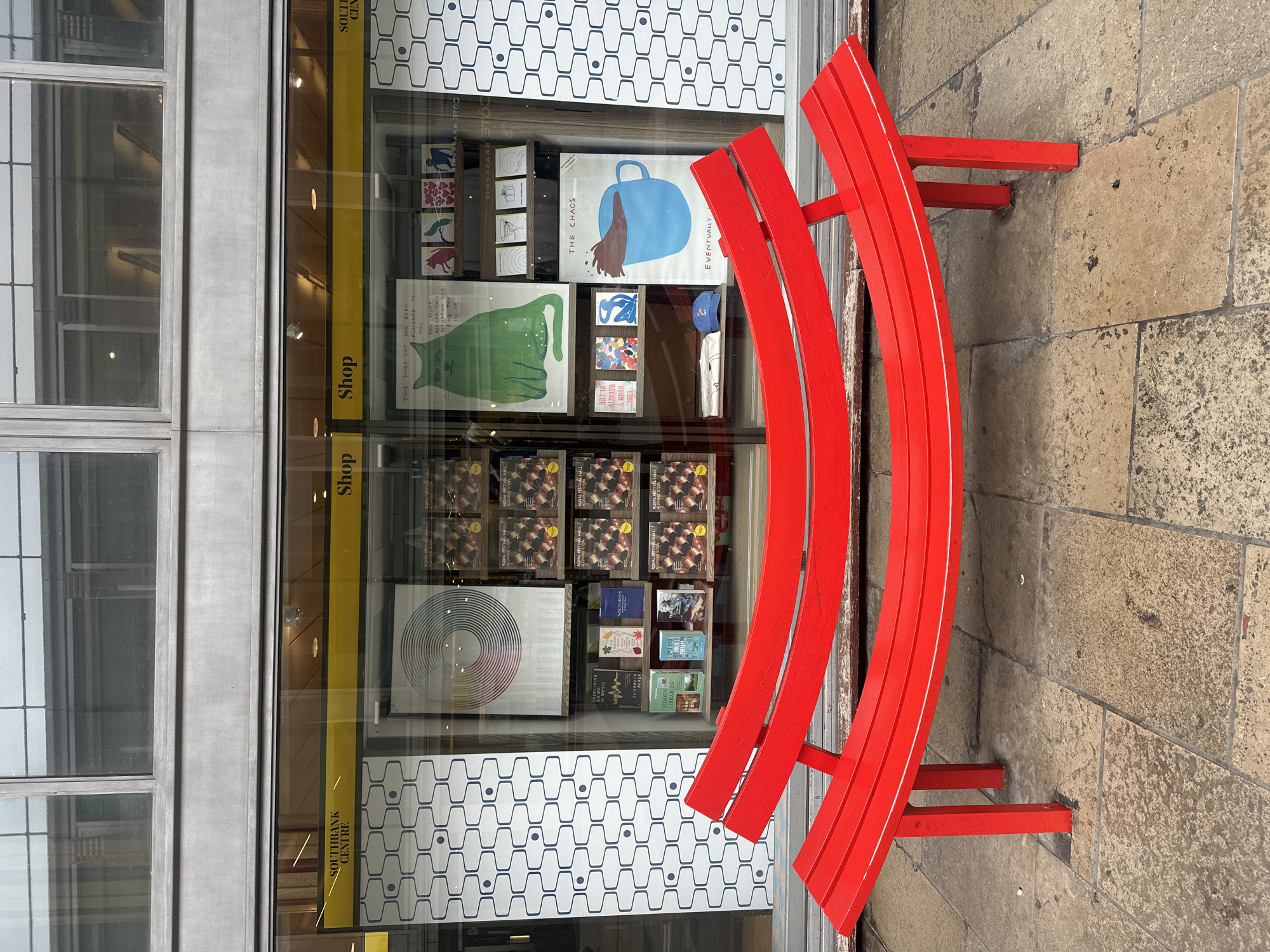 Here's a detailed description of the image:
The photograph shows the exterior of a shop located within the Southbank Centre in London, United Kingdom. The shop's window display is filled with posters, books, and other merchandise arranged on shelves and in display cases.
Several posters are prominently featured: one depicts a blue cup with brown liquid spilling from it, titled "The Chaos" and "Eventually". Another poster features a green cat and the text "This huge cat has been assigned to you. You must accept this situation and find joy in it". Other items include books with titles such as "Hidden Libraries", "Is Music", and "Eliza Alher". A design of concentric circles is also displayed.
In front of the shop window sits a curved, bright red bench. It is made of multiple curved wooden slats supported by a frame of the same color. The bench is located on a paved area with light brown stone tiles. The shop's window is partitioned into smaller glass sections framed in gray. Above the window is a larger glass area that reflects the buildings and sky opposite the shop.
The environment appears to be an urban, pedestrianized area within a cultural center. The overall impression is that of a bookshop selling art prints and related items within a notable arts venue in London.