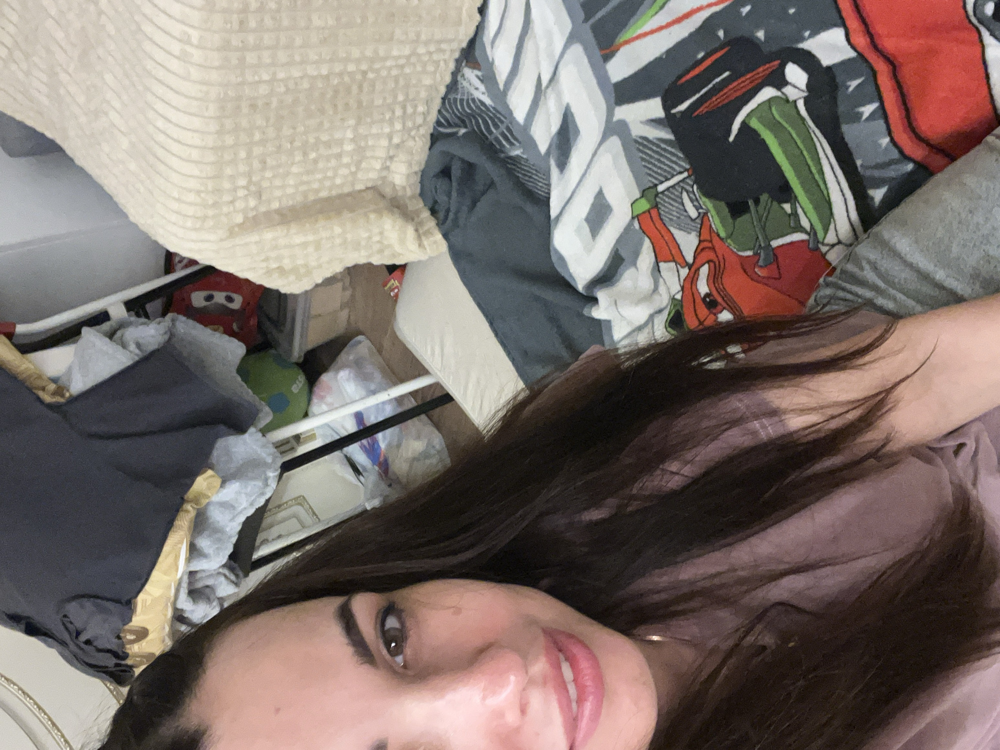 A woman with long dark hair and brown eyes is looking at the camera in a selfie. She is wearing a pink shirt and is sitting on a bed with a blanket that has a Cars movie theme. In the background, there are clothes piled up, a white chair, and a beige blanket on another bed.
