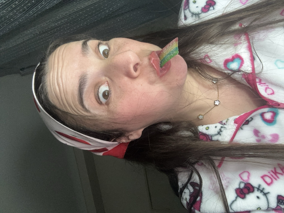 A woman lies sideways, staring wide-eyed. She wears a white headband with red accents, brown hair, and a pajama top with a Hello Kitty pattern. A rainbow-colored candy strip is held between her lips. A delicate gold necklace with clover-shaped pendants is visible on her neck. The background includes a textured gray curtain.