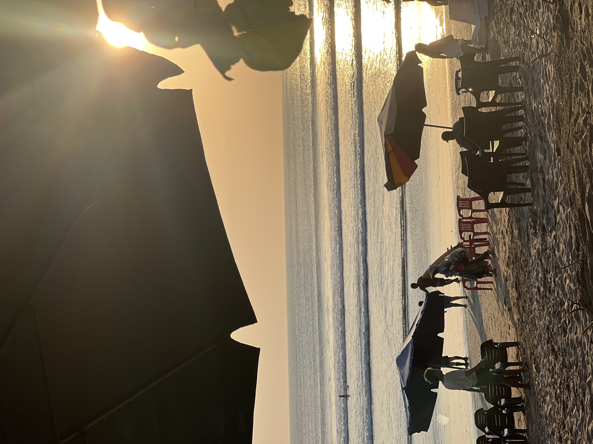This image depicts a sunset scene on Kuta Beach in Kuta, Indonesia. The foreground is dominated by sand and several beach chairs, some of which are arranged around small tables. Beach umbrellas are set up, providing shade.
In the middle ground, several people are visible on the sandy beach. Some appear to be walking or standing, while others are near the water's edge. One individual is seen standing on a surfboard in the ocean. Waves are breaking on the shore, and the ocean stretches out to the horizon.
The background is filled with the vast expanse of the ocean and the sky, illuminated by the warm glow of the setting sun. The sun is positioned high in the sky, with its rays creating a bright flare and reflecting off the water's surface. The overall atmosphere is serene, characteristic of a beach at dusk.