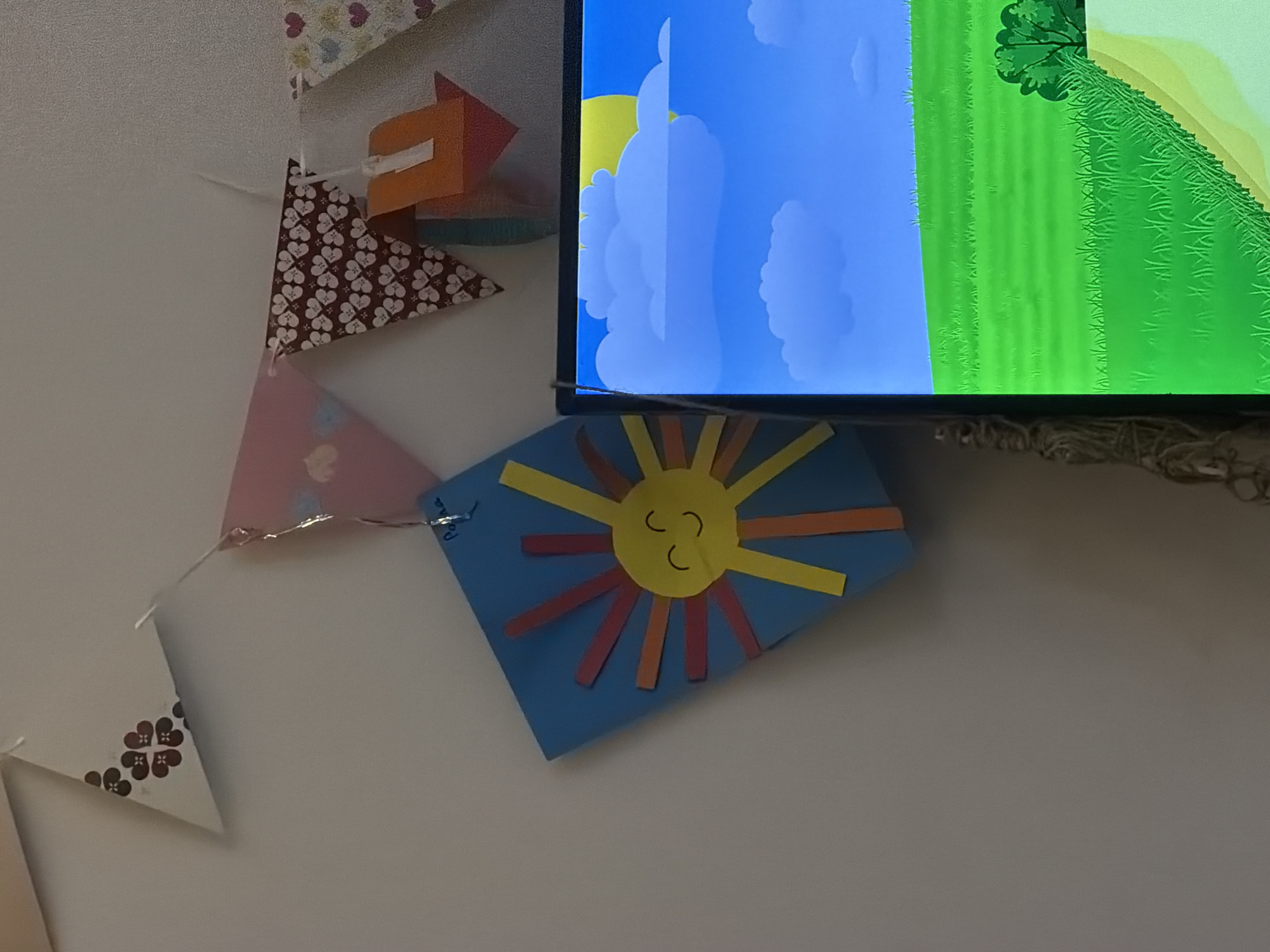 The image shows a wall with a string of triangular flags. On the left, a blue folded paper craft with a yellow sun and colorful rays is attached to the wall. To the right of the craft, a monitor displays a cartoon landscape with a bright blue sky, white clouds, and green fields. A thin wire hangs vertically on the left side of the monitor. The flags are decorated with patterns of hearts and flowers. No people are visible in the image. The location is not specified in the image.