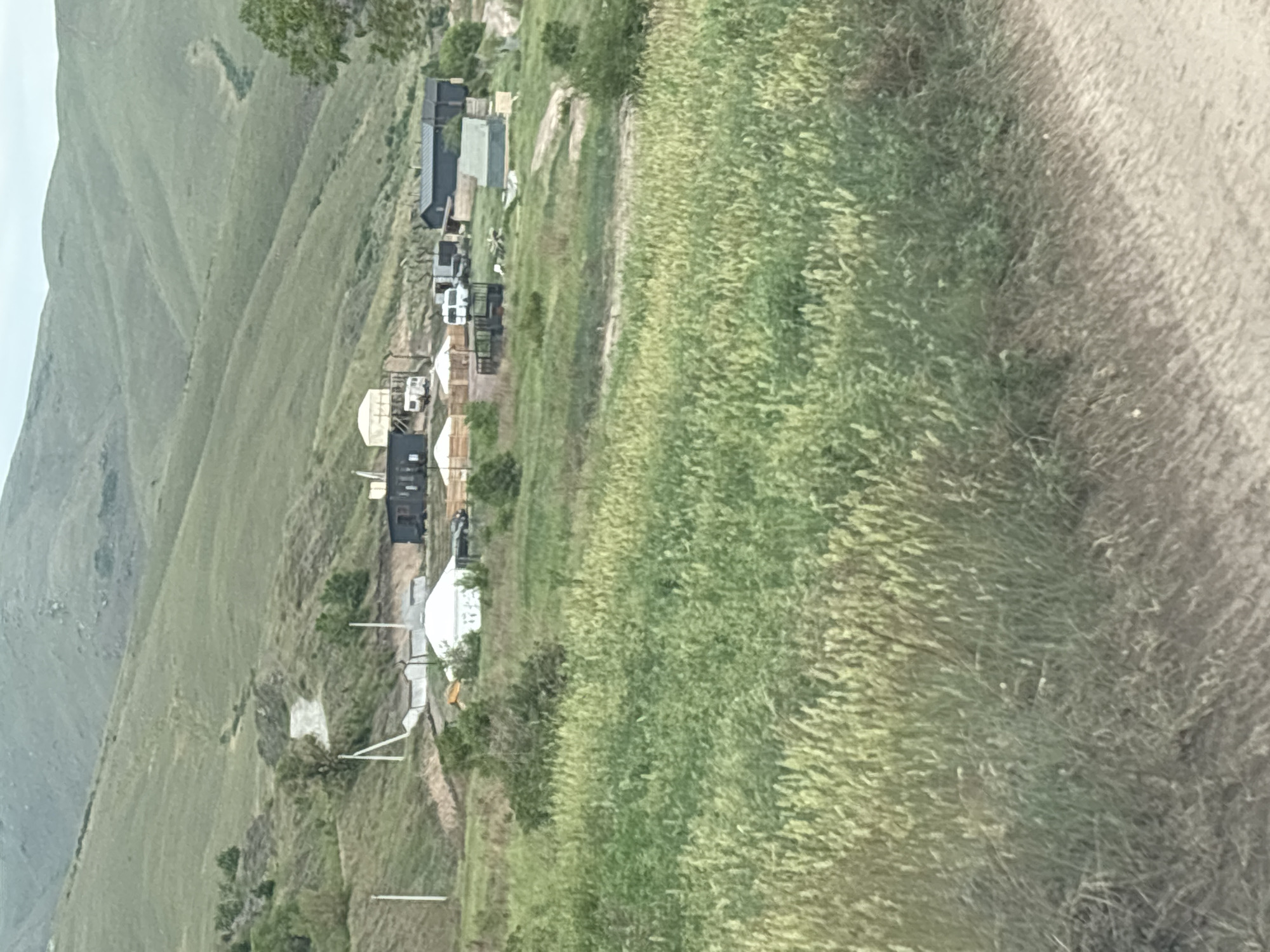 The image depicts a rural scene with several yurts and small buildings situated on a hillside in what appears to be a resort or nomadic settlement. The yurts are circular, tent-like structures with conical roofs, and some are white while others have darker exteriors. Several vehicles, including vans and what looks like a truck or utility vehicle, are parked near the structures. The surrounding environment consists of rolling green hills covered in grass and vegetation. A dirt road or pathway is visible in the foreground and winding up the hillside. There are no people visible in the image. The overall impression is one of a rustic, possibly eco-tourism or historical lodging facility. The location is identified as Ust-Abakan, Russia.