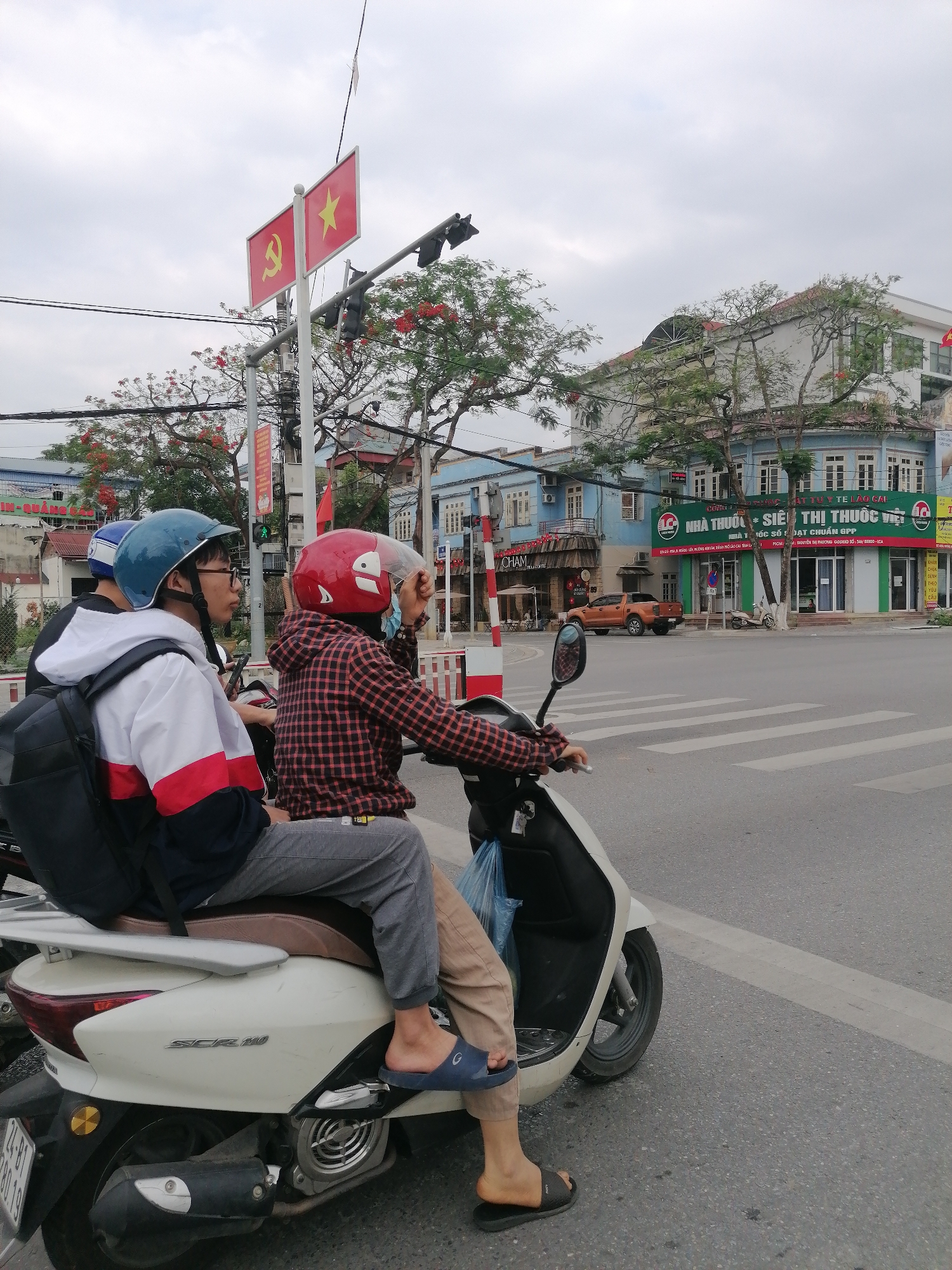 Two individuals on a white scooter wait at a traffic light in Lao Cai Ward, Vietnam. The driver wears a red plaid shirt and red helmet, while the passenger sports a blue helmet and a jacket with white, red, and navy sections. A Vietnamese flag is visible near the traffic light. Buildings line the street, including a pharmacy and a café, with an orange pickup truck parked nearby.