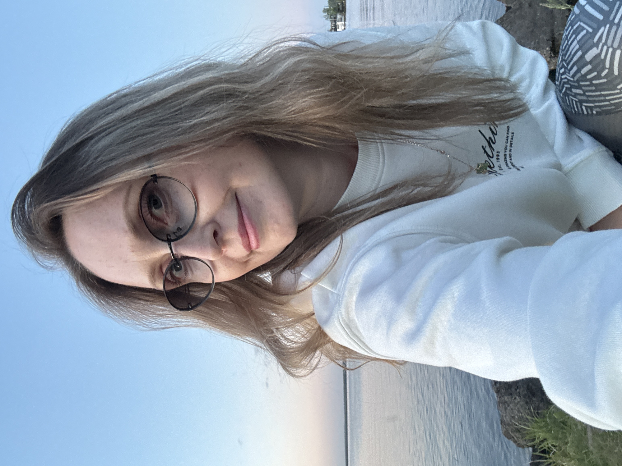 A woman with long blonde hair and wearing glasses poses for a selfie. She is wearing a white sweatshirt and a necklace. The background shows a body of water, likely a river or lake, and a hint of green vegetation. The sky is light blue, suggesting it is either early morning or late afternoon. The exact location in Chaikovsky, Russia, is not discernible from the image.