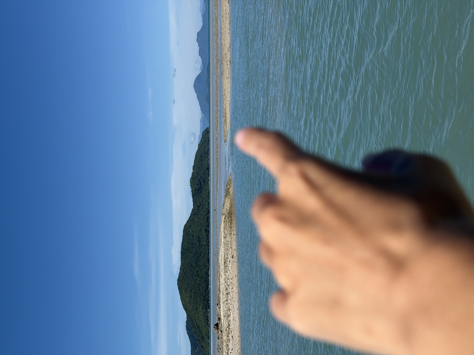 The image depicts a scenic coastal view in Baan Thong Krut, Thailand, dominated by a clear blue sky and turquoise waters. In the foreground, a blurred human hand with an outstretched index finger points towards the horizon. The middle ground features a sandy island with scattered rocks, partially submerged in the shallow, rippling water. Beyond the island, lush green hills and mountains rise, forming a picturesque backdrop. The overall scene captures a serene and natural environment, possibly during daylight hours given the bright illumination.