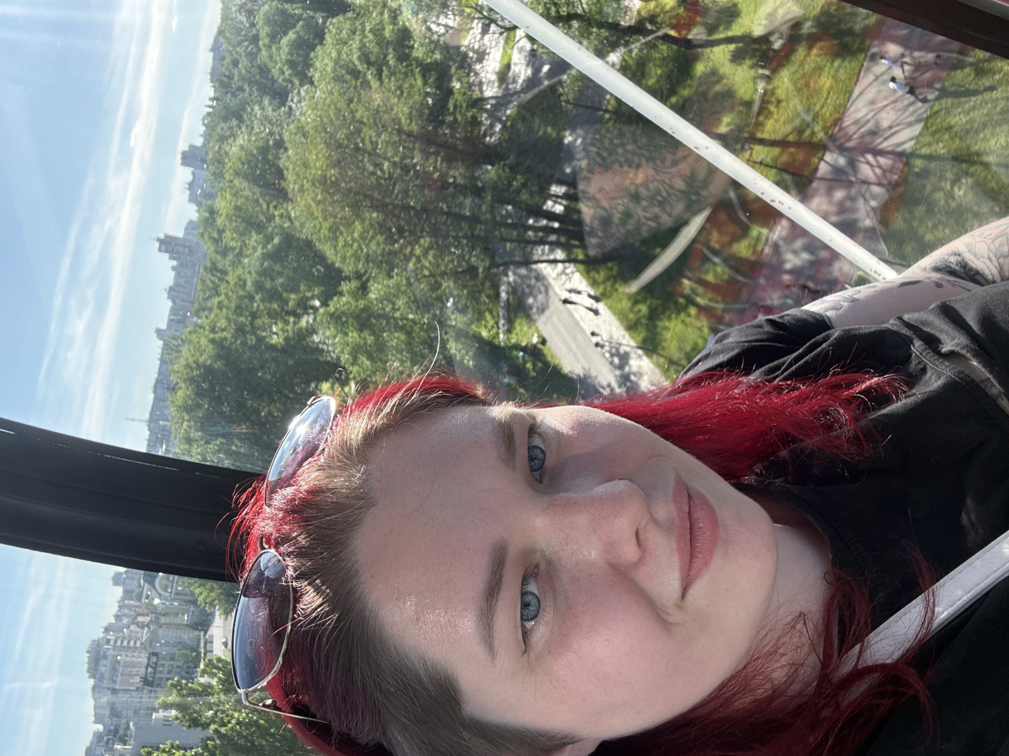 The image presents a selfie-style portrait of a person with dyed red hair and blue eyes, taken from inside an elevated structure, overlooking the city of Lipetsk, Russia. The individual, positioned in the lower-left quadrant, has light skin, a subtle smile, and a small ring piercing the left nostril. Their hair is predominantly red and auburn, with one side appearing shorter or shaved, and a pair of dark sunglasses rests on top of their head. They are wearing a black short-sleeved top, revealing a portion of tattoos on the upper right arm and shoulder. A white strap is visible across their left shoulder.
Through the window of the structure, a vast outdoor scene is visible under a clear blue sky with minimal cloud cover. The immediate background shows an expansive green park or forested area, characterized by numerous deciduous trees. Paved pathways are discernible within the park, with several figures appearing as silhouettes walking along them. Beyond the park, an urban landscape extends, comprising various multi-story buildings, indicating a developed city environment. Parts of the internal structure, including a prominent black vertical support beam on the right and a white diagonal bar extending from the lower right, are visible, suggesting the person is inside a gondola, cable car, or observation pod. Reflections of the exterior scene are present on the glass surface.