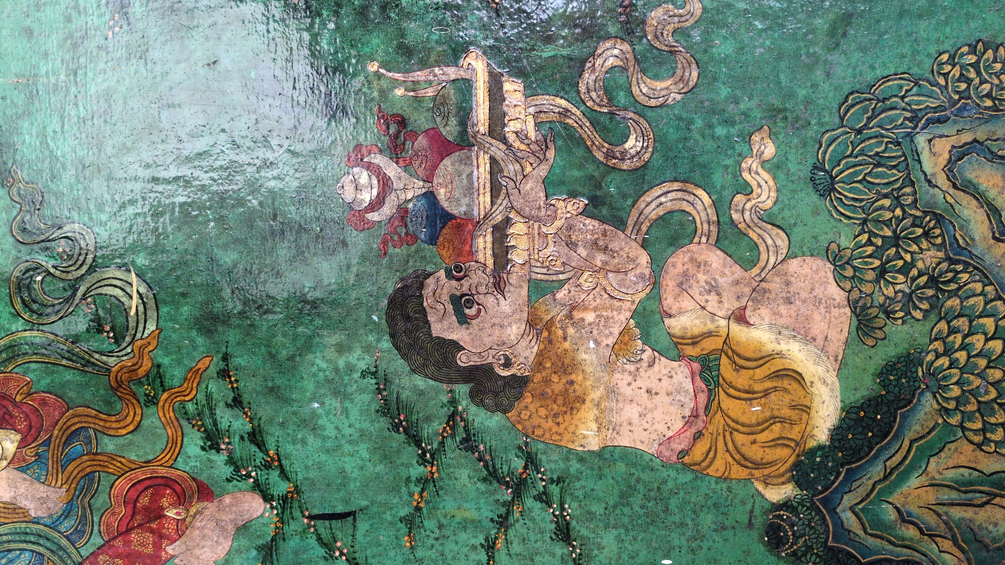 The image shows a vibrant, heavily stylized artwork, likely a mural, painted on a surface with a weathered, textured, green background. The artwork depicts a figure hanging upside down, grasping a vertical object topped with a cluster of colored spheres. The figure has tan skin, dark hair, and prominent eyes, wearing a yellow garment. Elaborate, stylized floral and foliate designs surround the figure, along with swirling patterns resembling ribbons or smoke. There is no identifiable location in the city of Fatehpur, India, discernible from the image alone.
